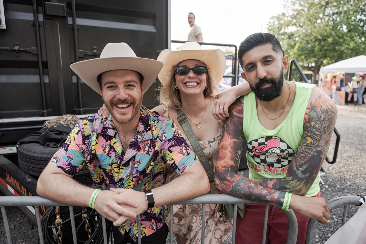 Three friends smile together at an outdoor festival, two wearing wide-brim hats, one with colorful tattoos.