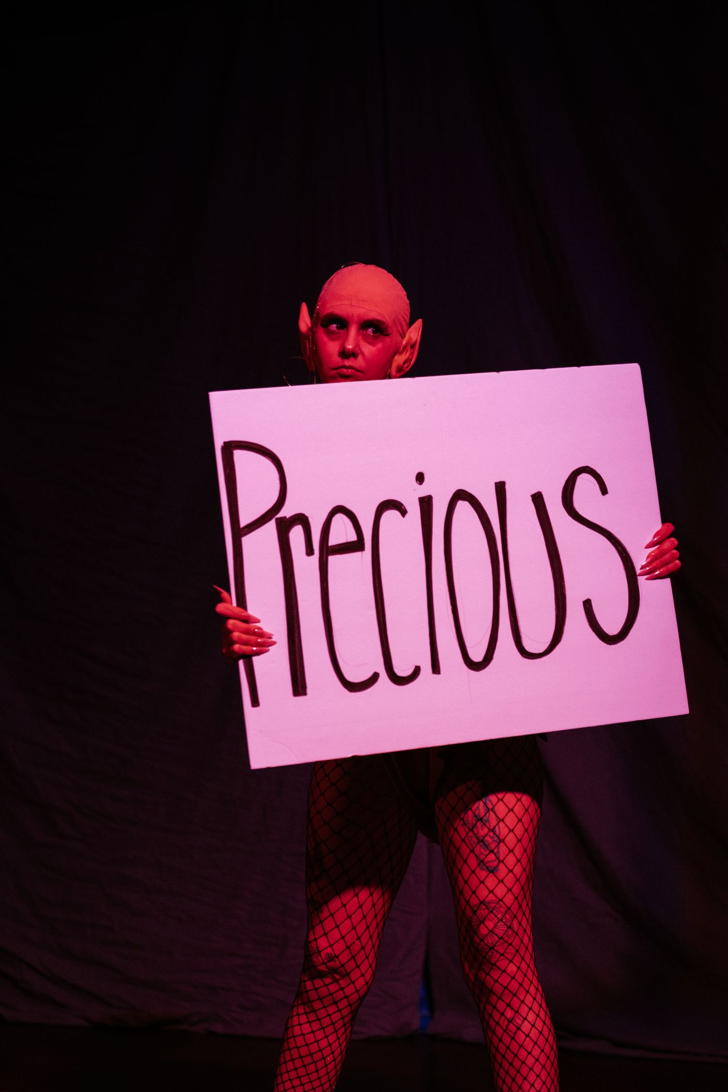 Bald performer in red lighting holds a pink sign reading Precious, wearing fishnet stockings on a dark stage.