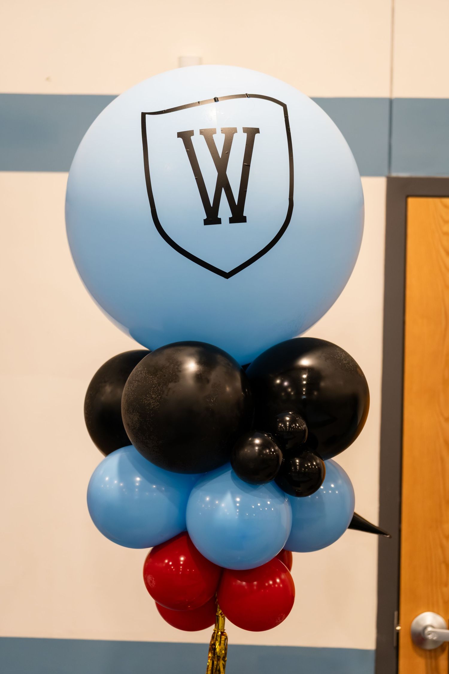 Blue balloon with W shield logo atop black, blue, and red balloon bouquet arrangement at school event.