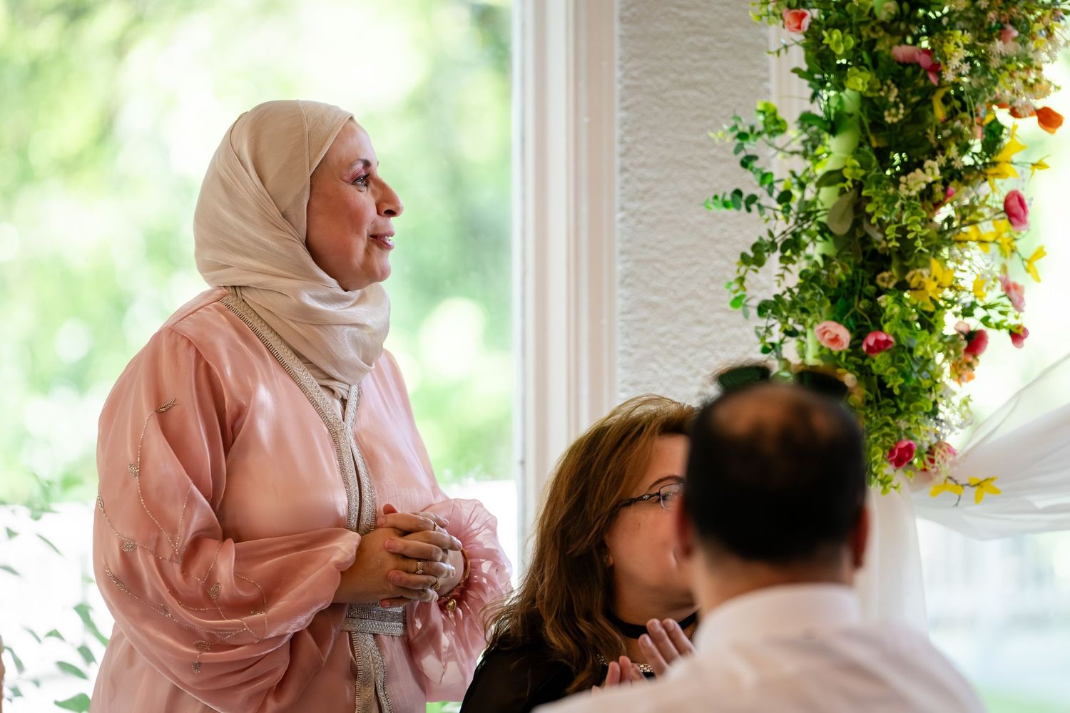 Woman in pink hijab stands near floral arrangement at elegant indoor gathering with guests seated nearby.