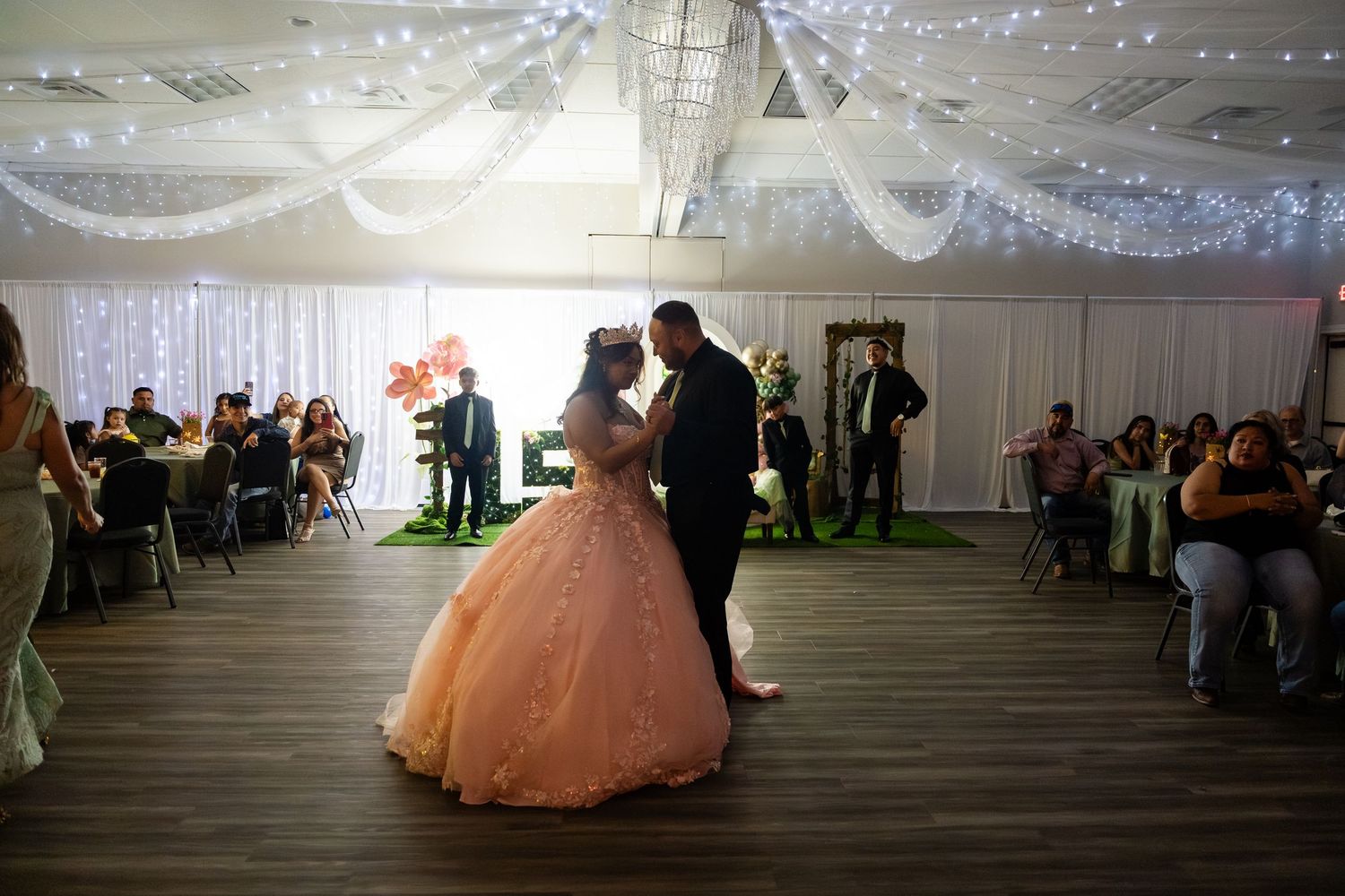 Couple sharing first dance at quinceañera celebration in elegant venue with fairy lights and white draping.