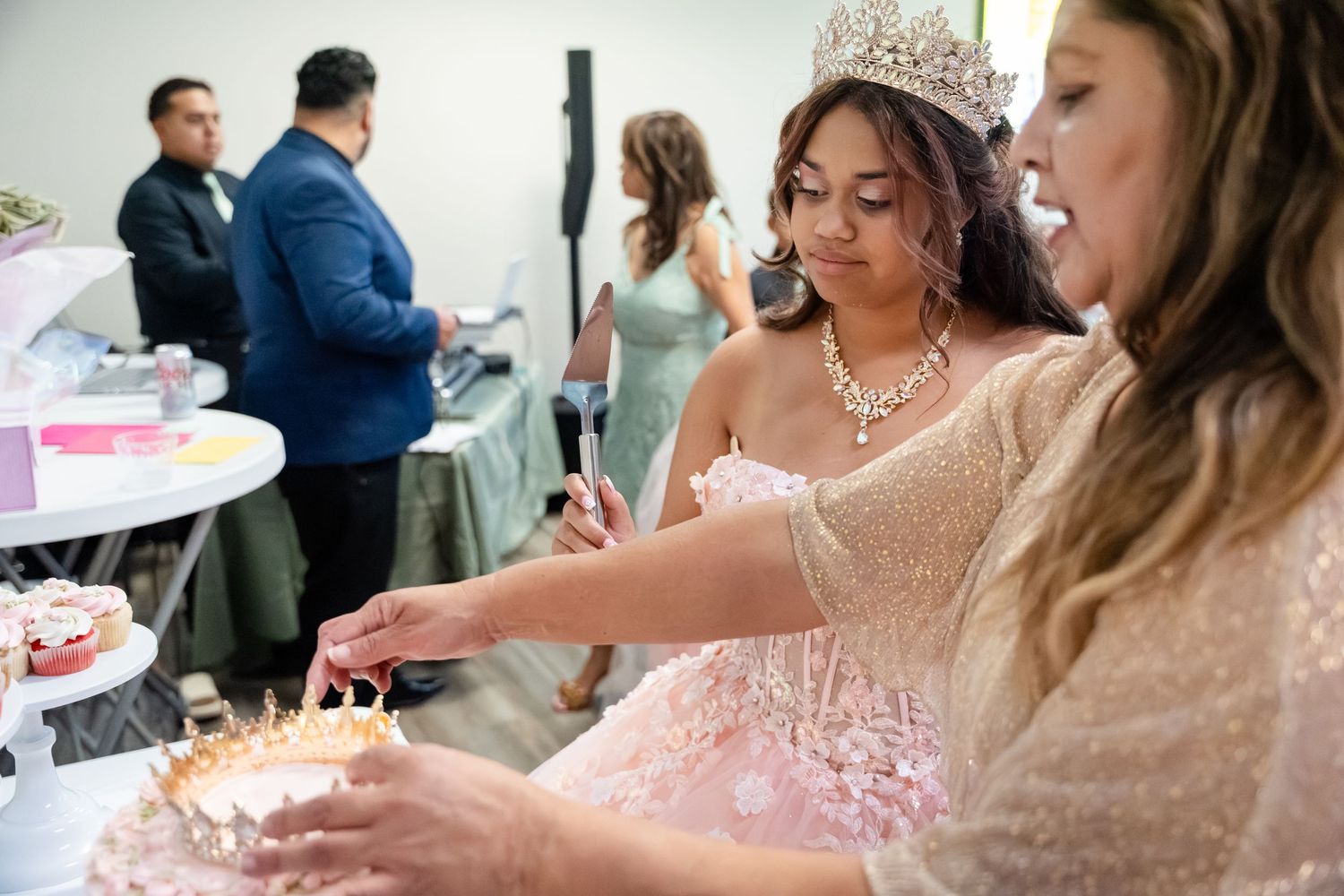 Young woman in pink quinceañera gown and tiara cuts cake at her celebration party with guests in background.