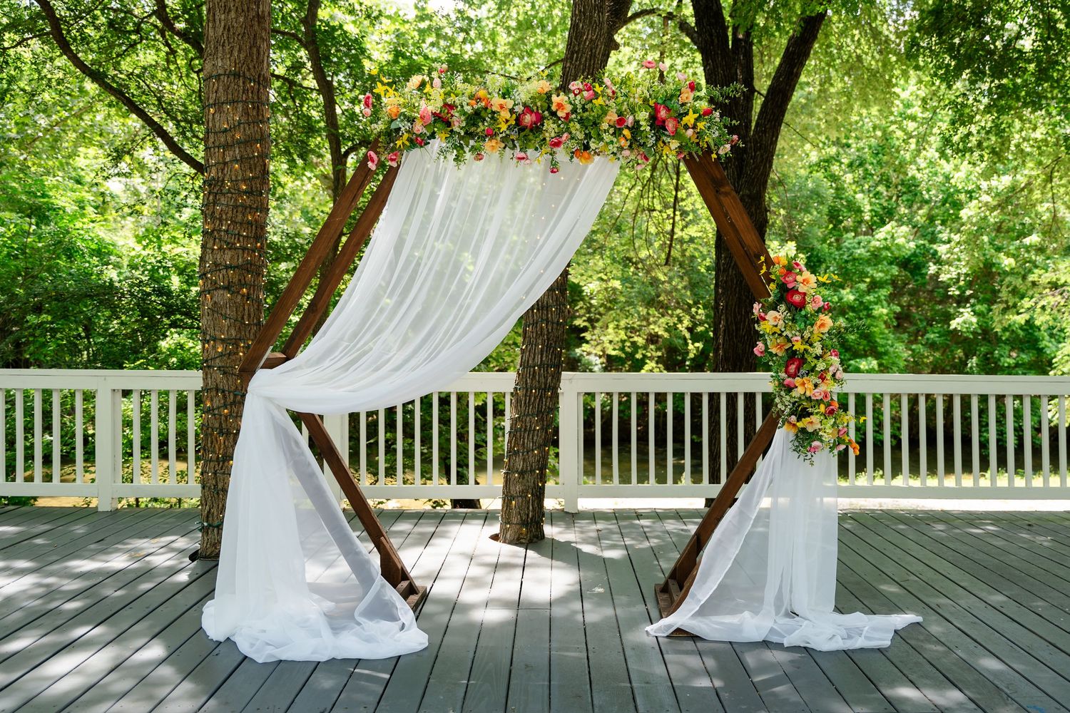 Outdoor wedding arch adorned with white draping and colorful flowers on a wooden deck surrounded by lush green trees.