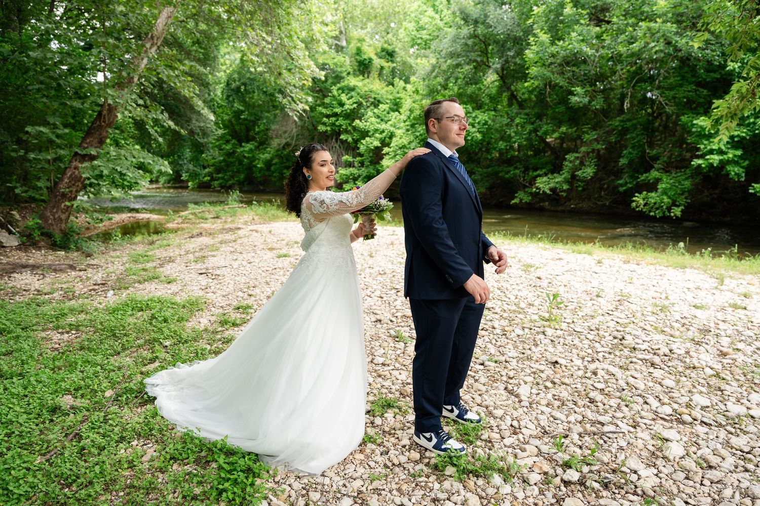 A bride in a white gown and groom in a navy suit dance outdoors near a rocky riverbank surrounded by lush green trees.