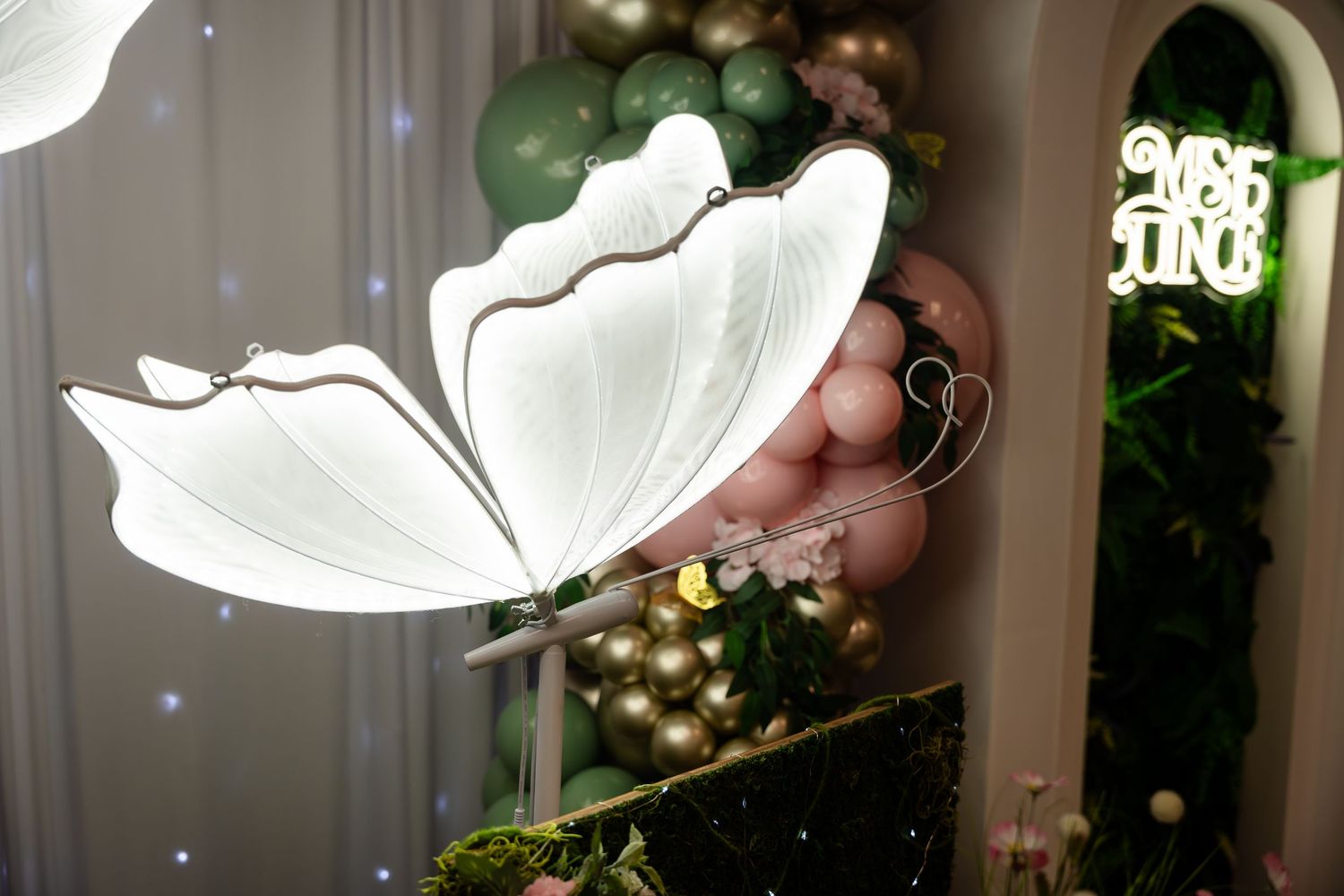 Large white butterfly decoration with glowing wings at an elegant event with green and gold balloon backdrop.