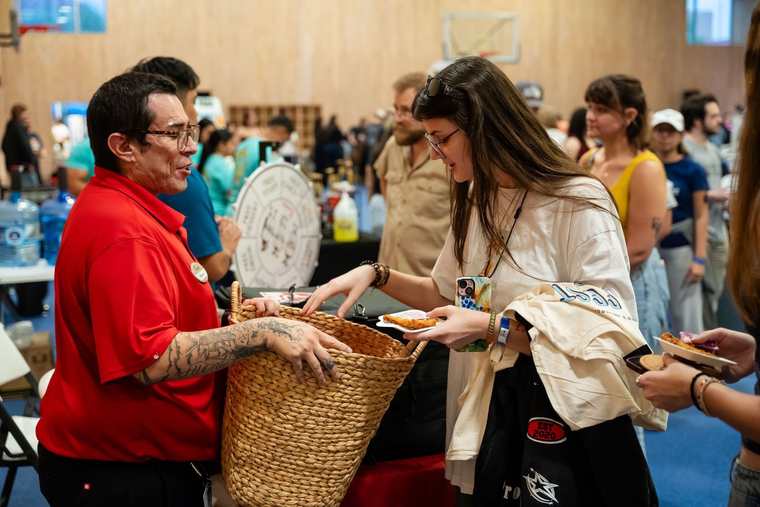 Two women exchange items at a busy indoor market event, one holding a woven basket at a community fair.