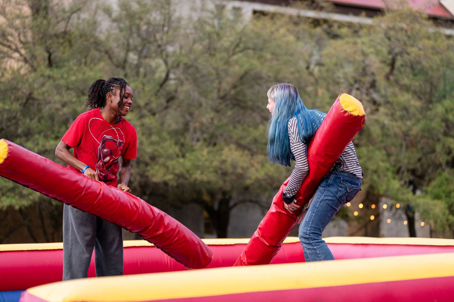 Two people playing inflatable jousting game at outdoor carnival event on colorful bouncy arena.