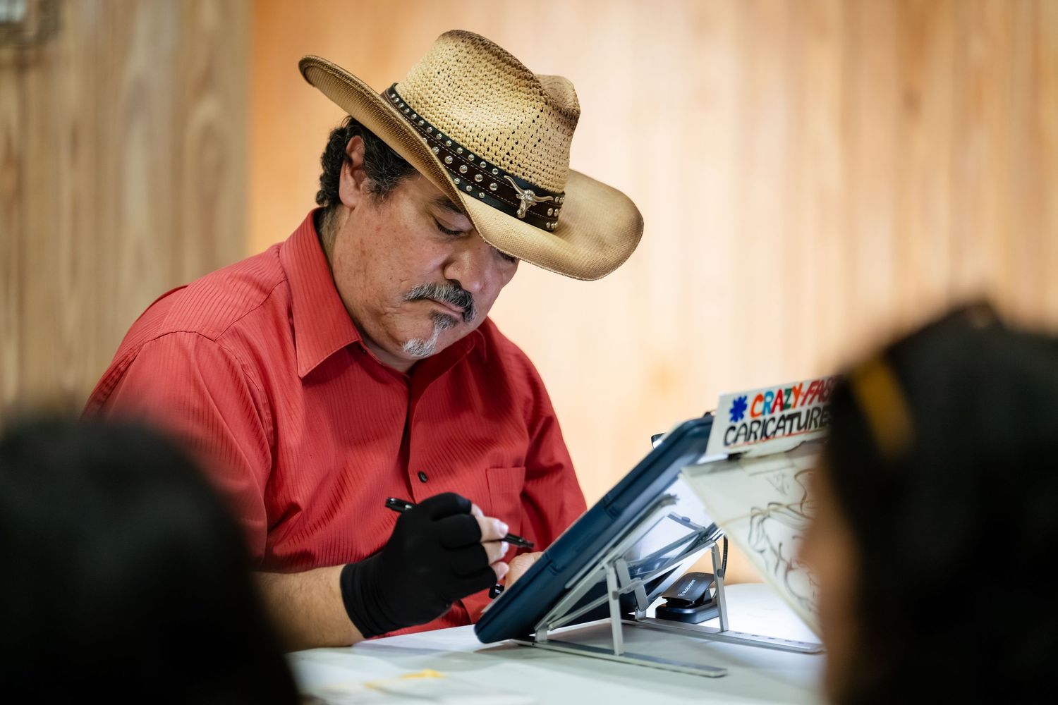 Man in straw cowboy hat and red shirt drawing on a tablet with a stylus at a table.
