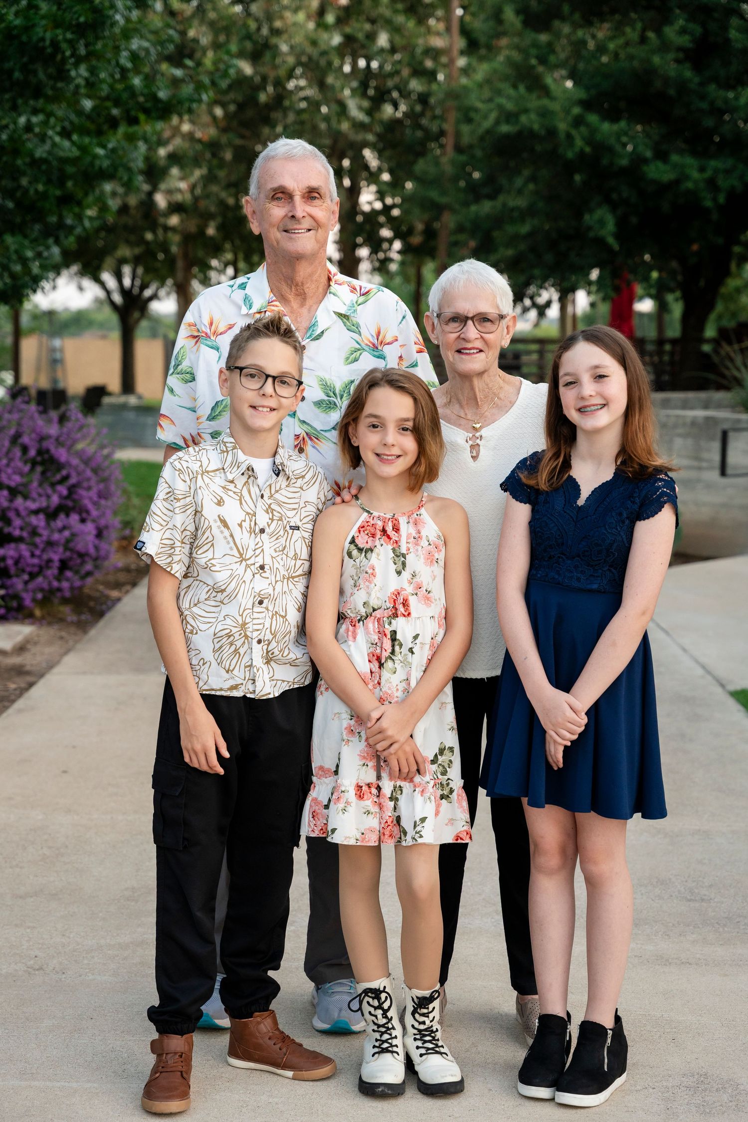 A family group poses together outdoors on a walkway with trees and flowers in the background.