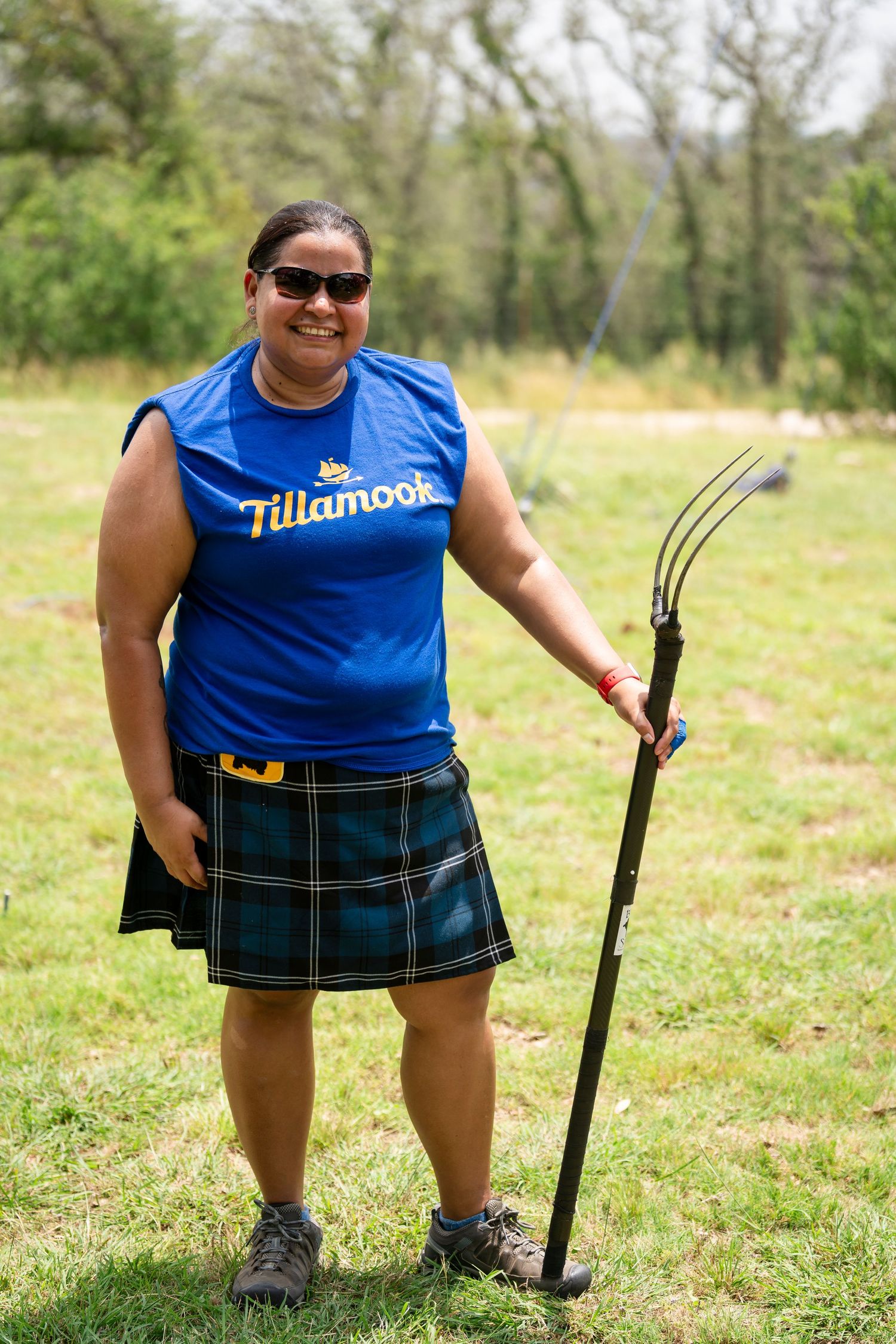 Someone wearing a blue t-shirt and plaid kilt stands in a grassy field holding a walking stick.
