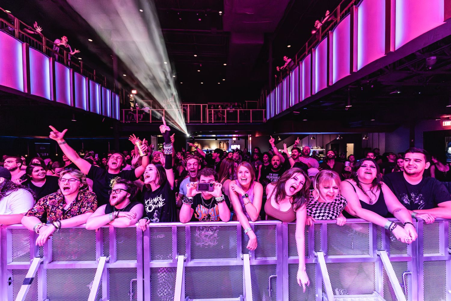 Excited concert crowd reaches toward stage barrier under purple venue lighting.