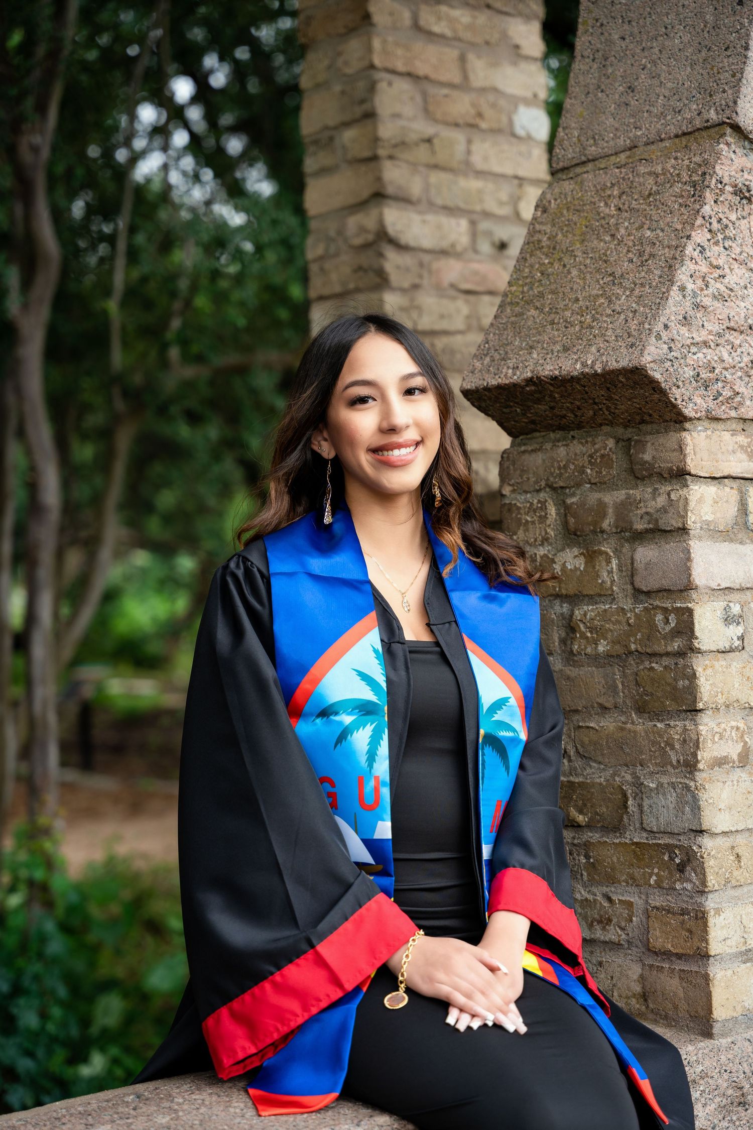 Graduation portrait featuring academic regalia with decorative blue and red stole against brick wall.