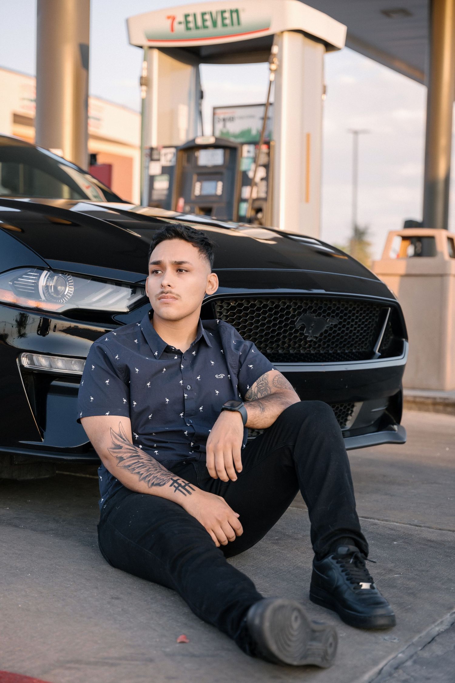 Person with tattoos sitting by black Mustang outside 7-Eleven gas station.