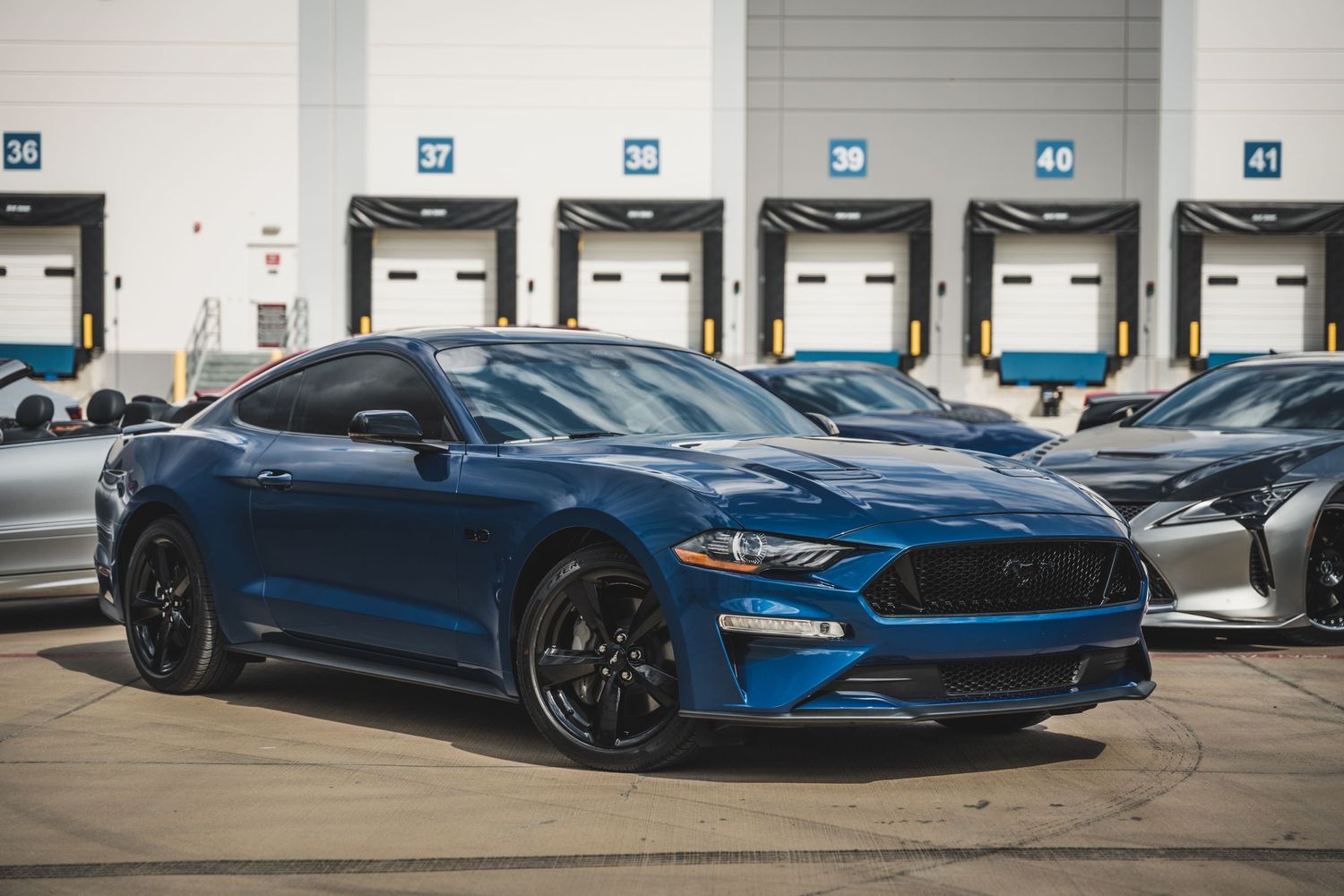 Blue Ford Mustang sports car with black wheels parked in front of warehouse loading docks.