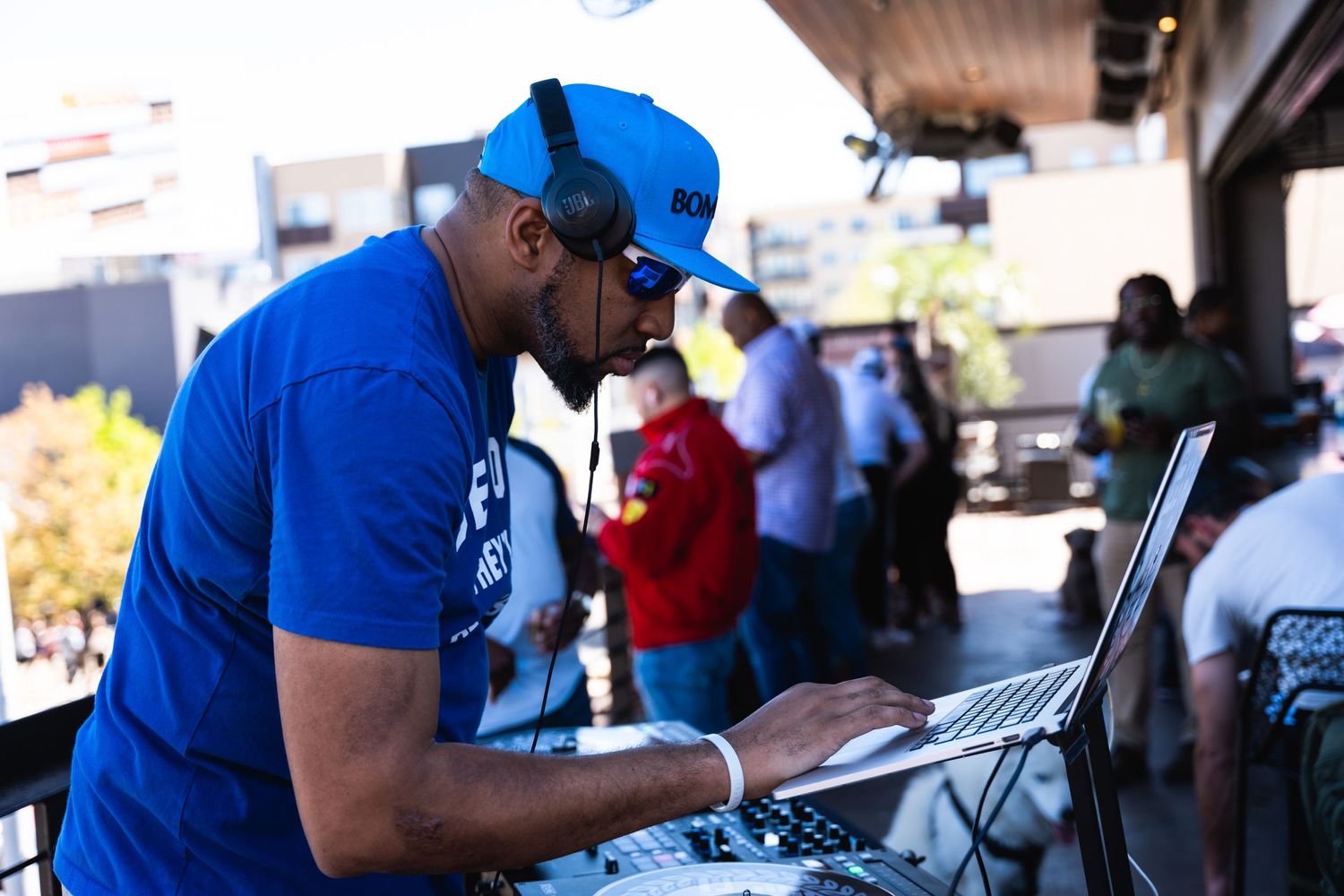 DJ in blue shirt and cap works on laptop equipment at outdoor event.