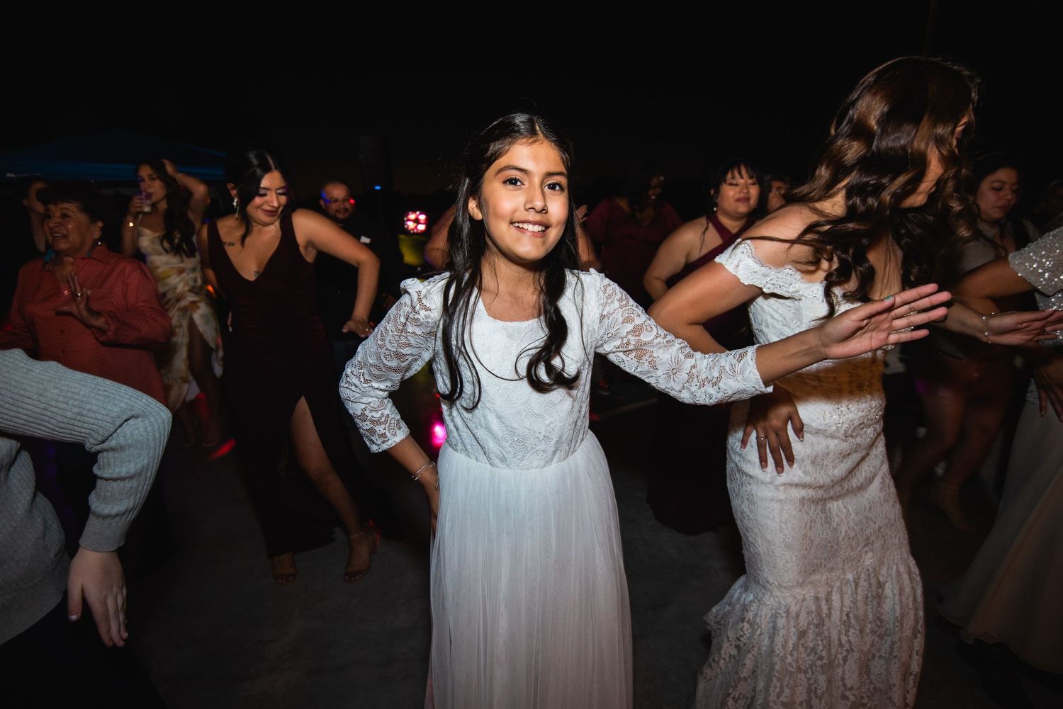 A young girl in a white dress dances joyfully at a celebration with other party guests in the background.