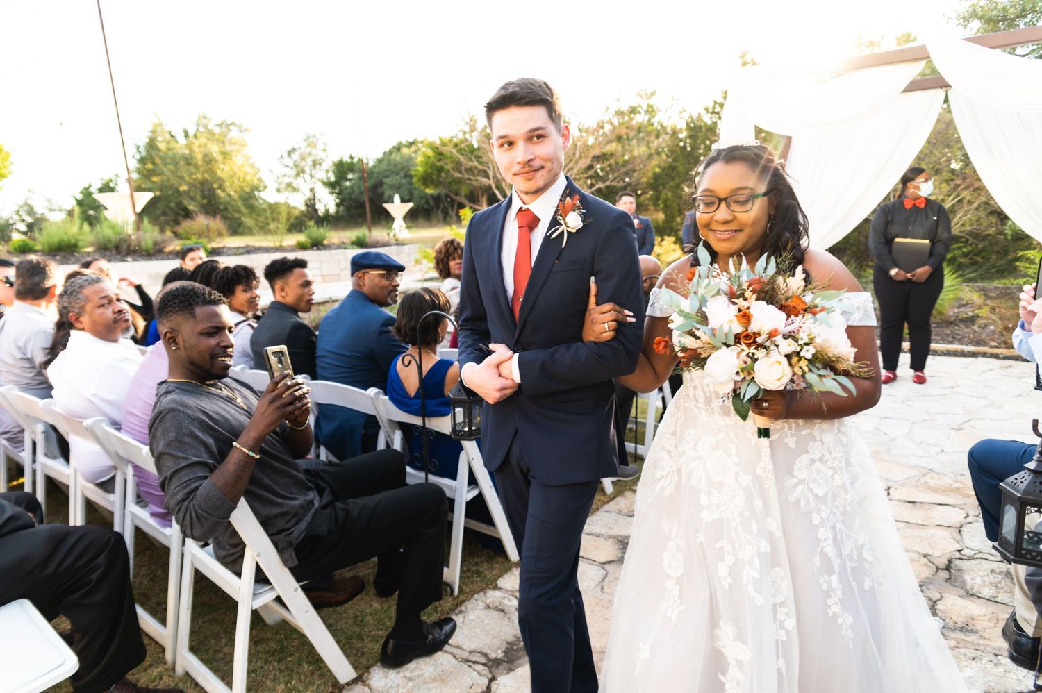 The newlyweds walking down the aisle as guests capture photos during the outdoor wedding recessional.