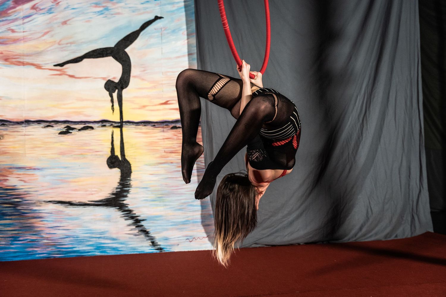 Aerial performer suspended from red silk ribbon against dramatic sunset backdrop during artistic performance.