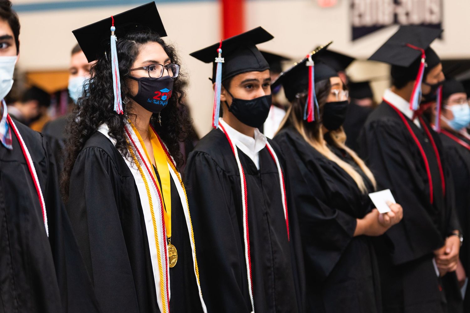 Row of graduates in black gowns and face masks at commencement ceremony.