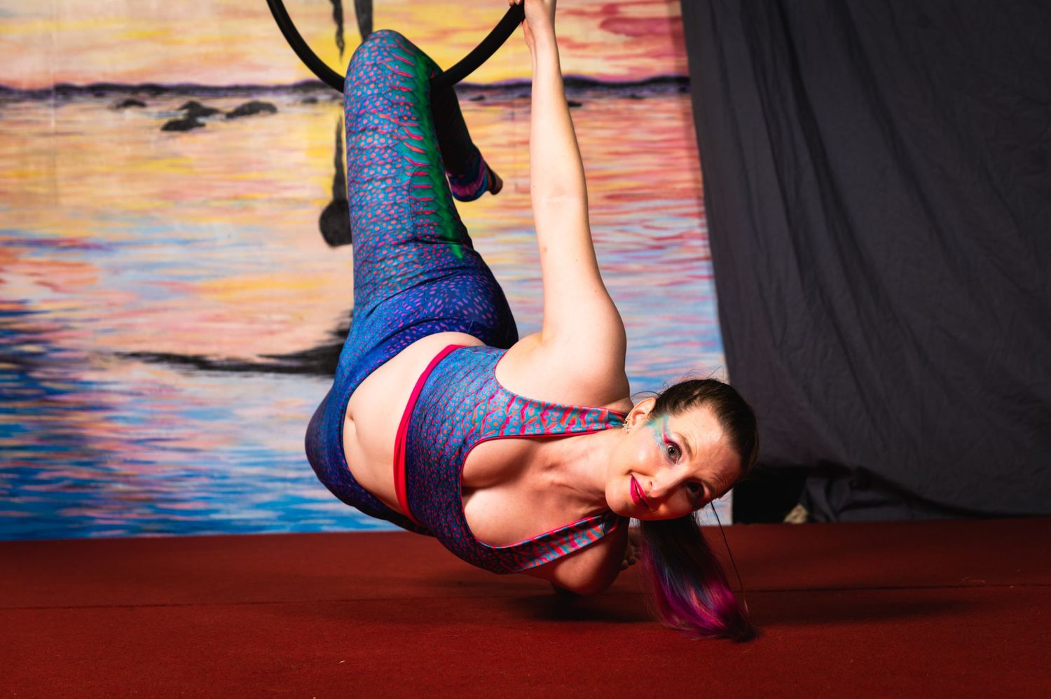 Aerial artist in iridescent blue costume performs on lyra hoop with sunset background.
