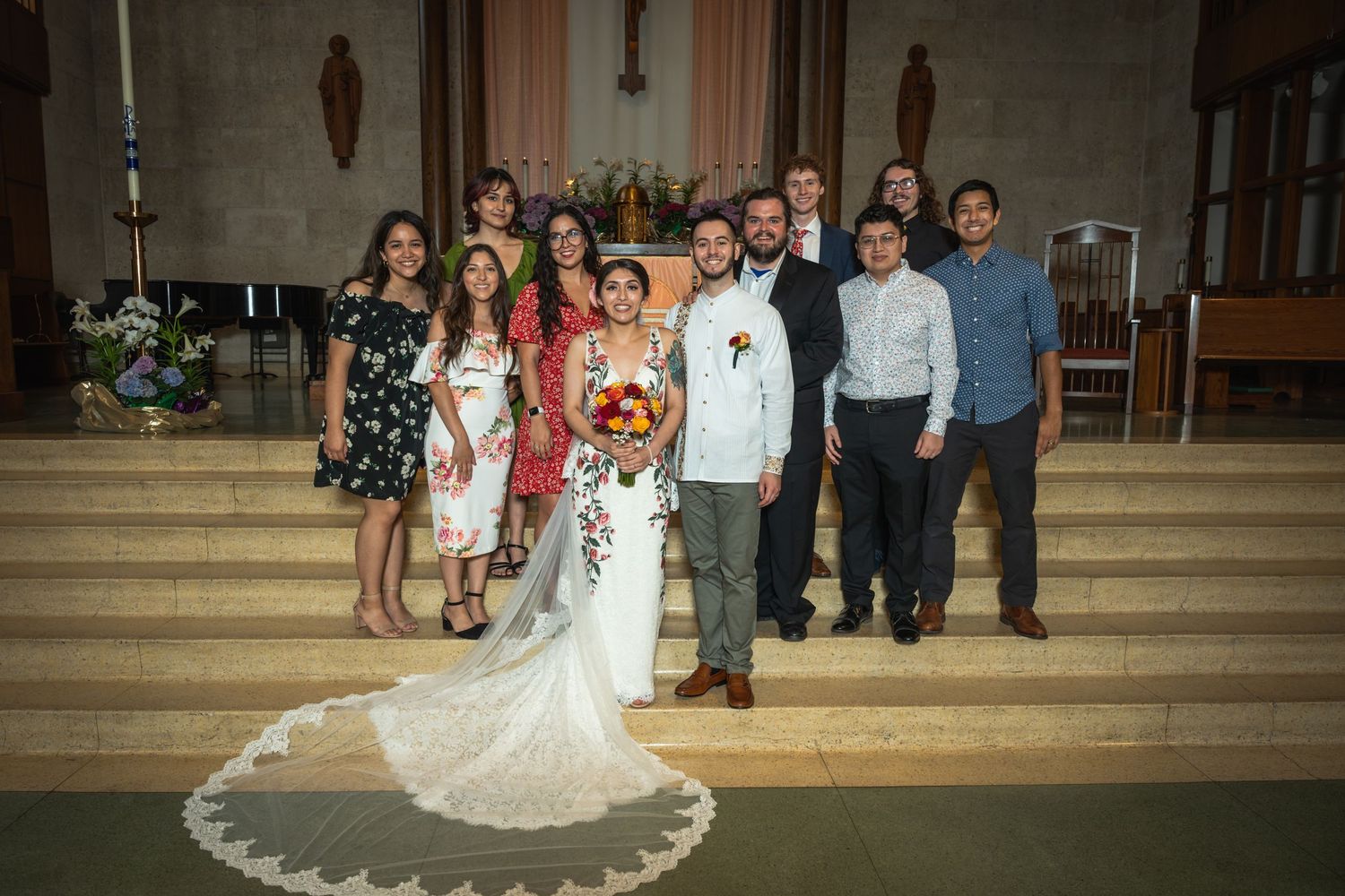 A wedding party poses for a group photo on church steps with the bride in a flowing white gown.