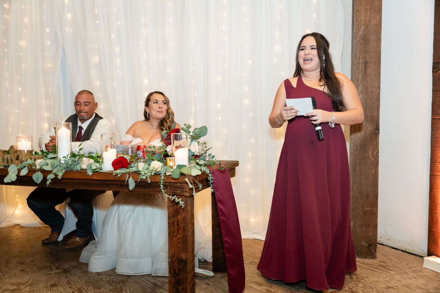 Wedding reception head table decorated with greenery and candles as guests listen to speeches.