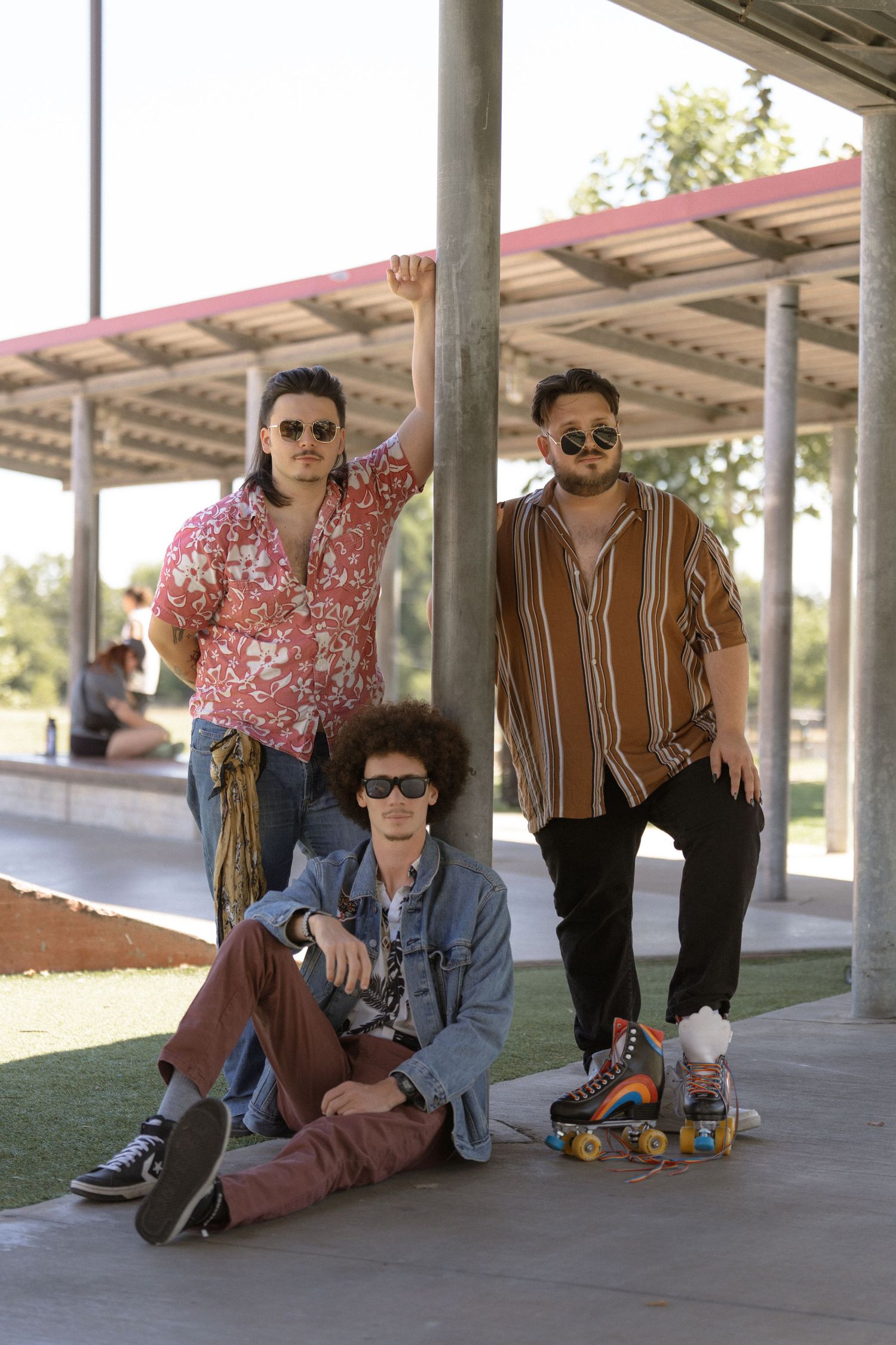 Three friends in retro-styled clothing pose with roller skates under a covered area.