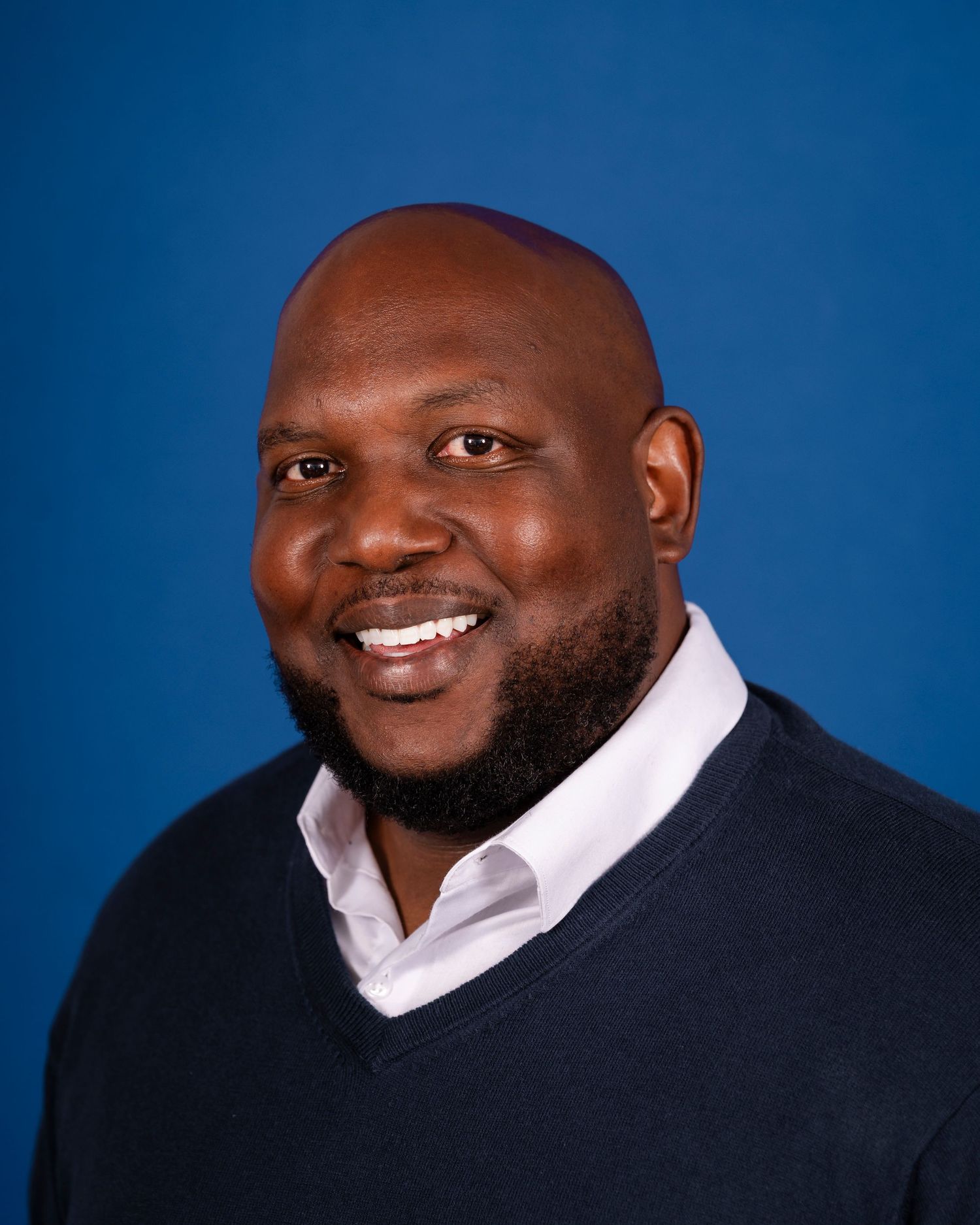 Professional headshot of person wearing navy sweater and white dress shirt against vivid blue background, smiling warmly.