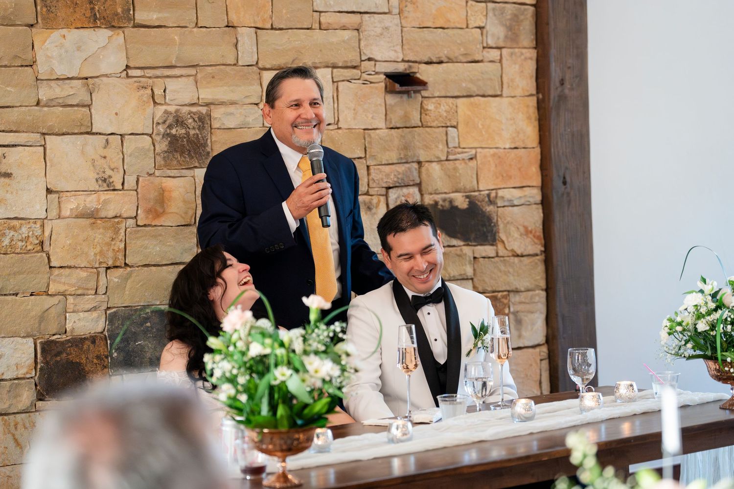 Wedding guests sharing laughter at a reception dinner with stone wall backdrop and elegant table settings.