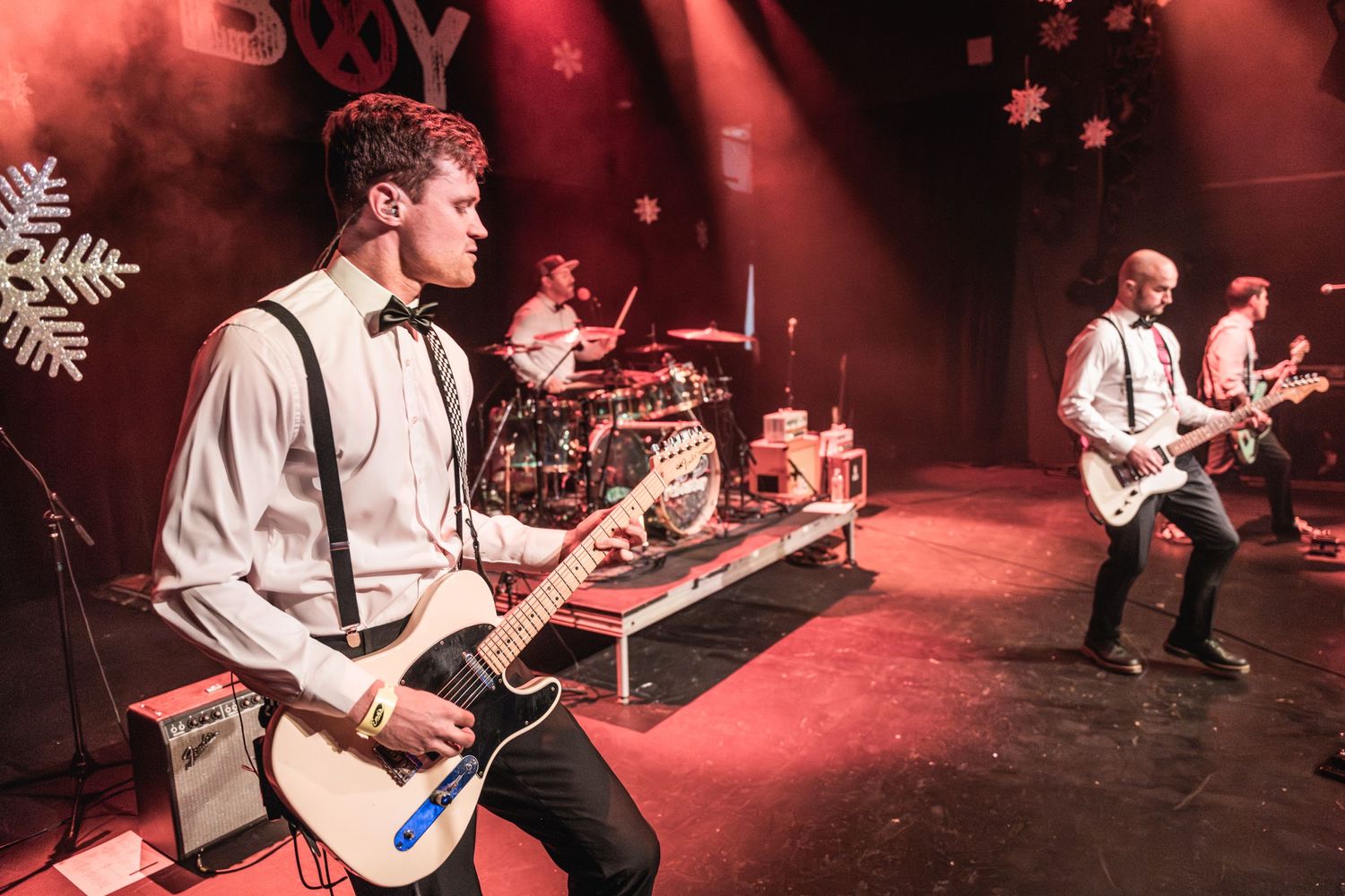 Band members in formal attire performing energetically on stage with red lighting and snowflake decorations.