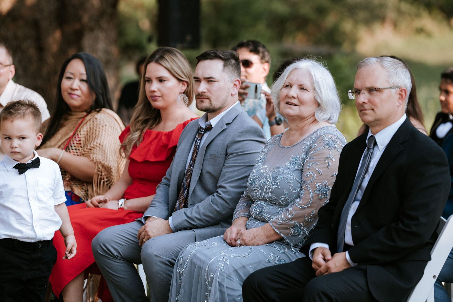 Wedding guests in formal attire seated on white chairs at an outdoor ceremony.