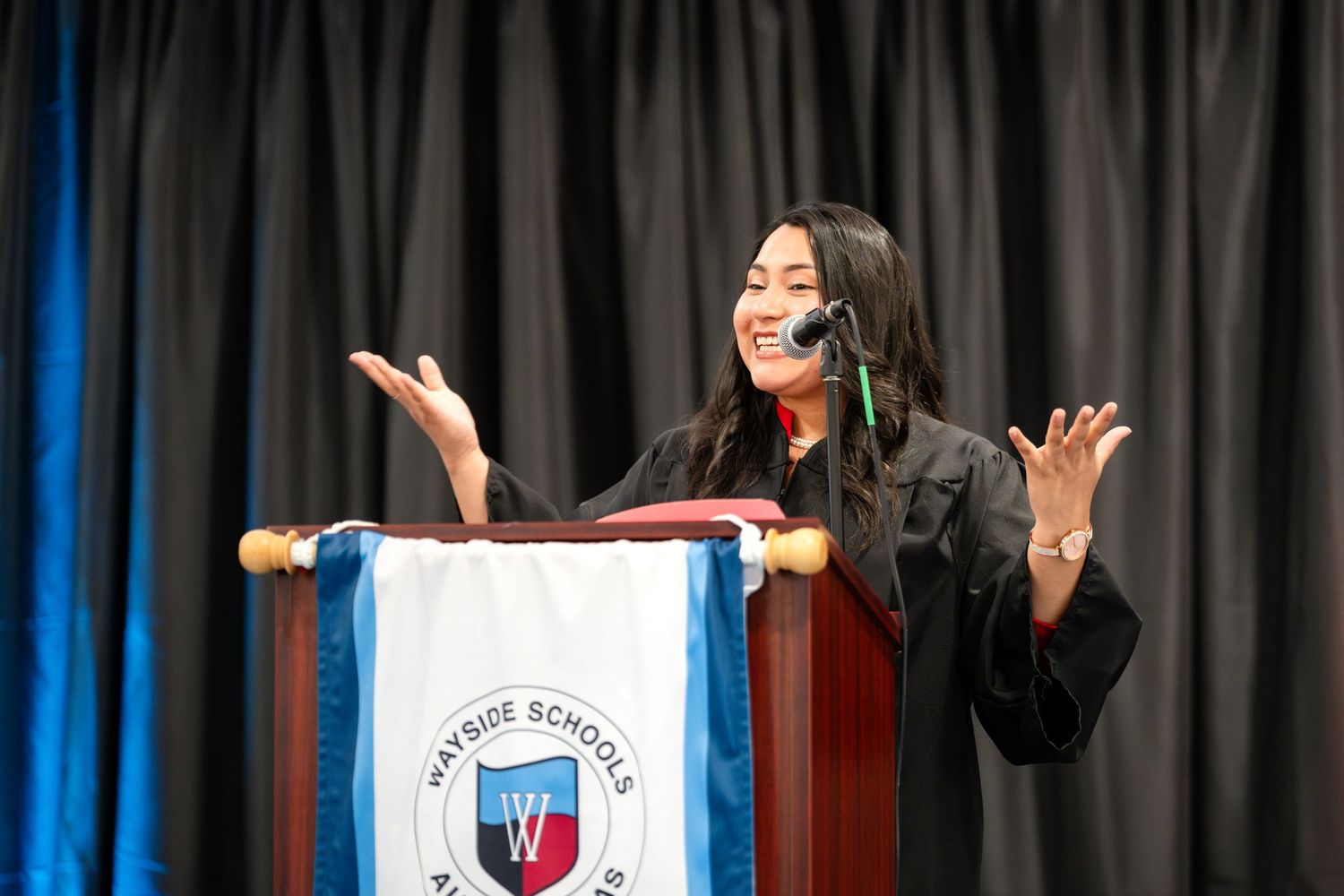 Graduate giving speech at podium with expressive hand gestures during ceremony.