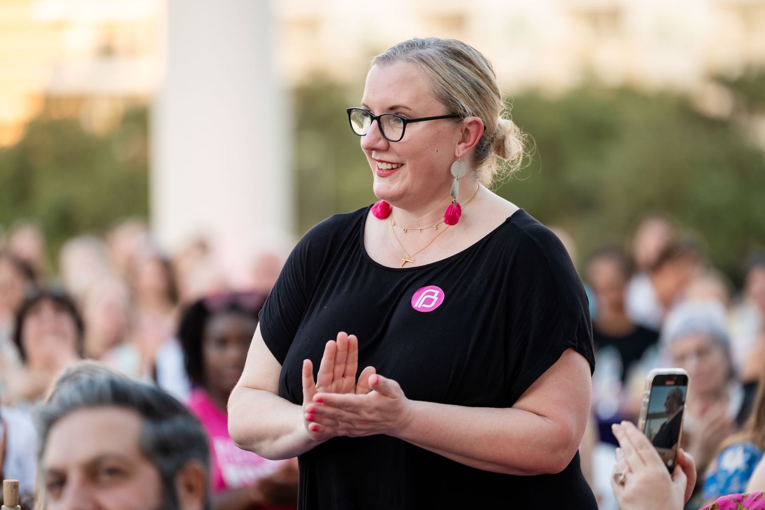Attendee in black shirt with pink button applauding during outdoor gathering.