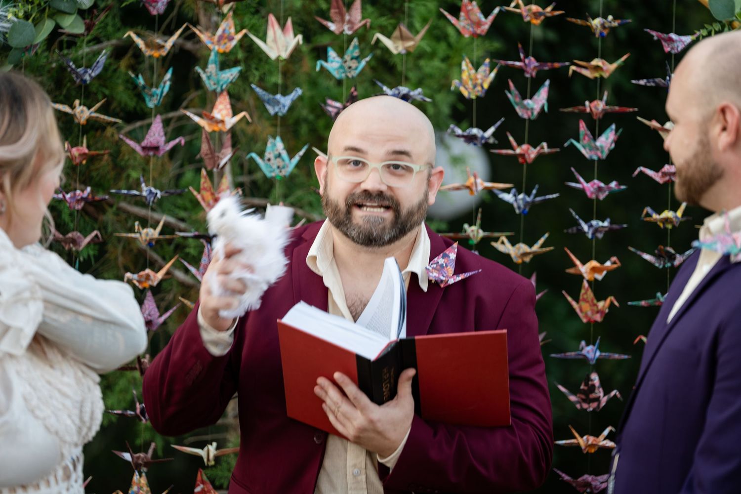 Officiant in burgundy suit stands before colorful paper crane backdrop during wedding ceremony.
