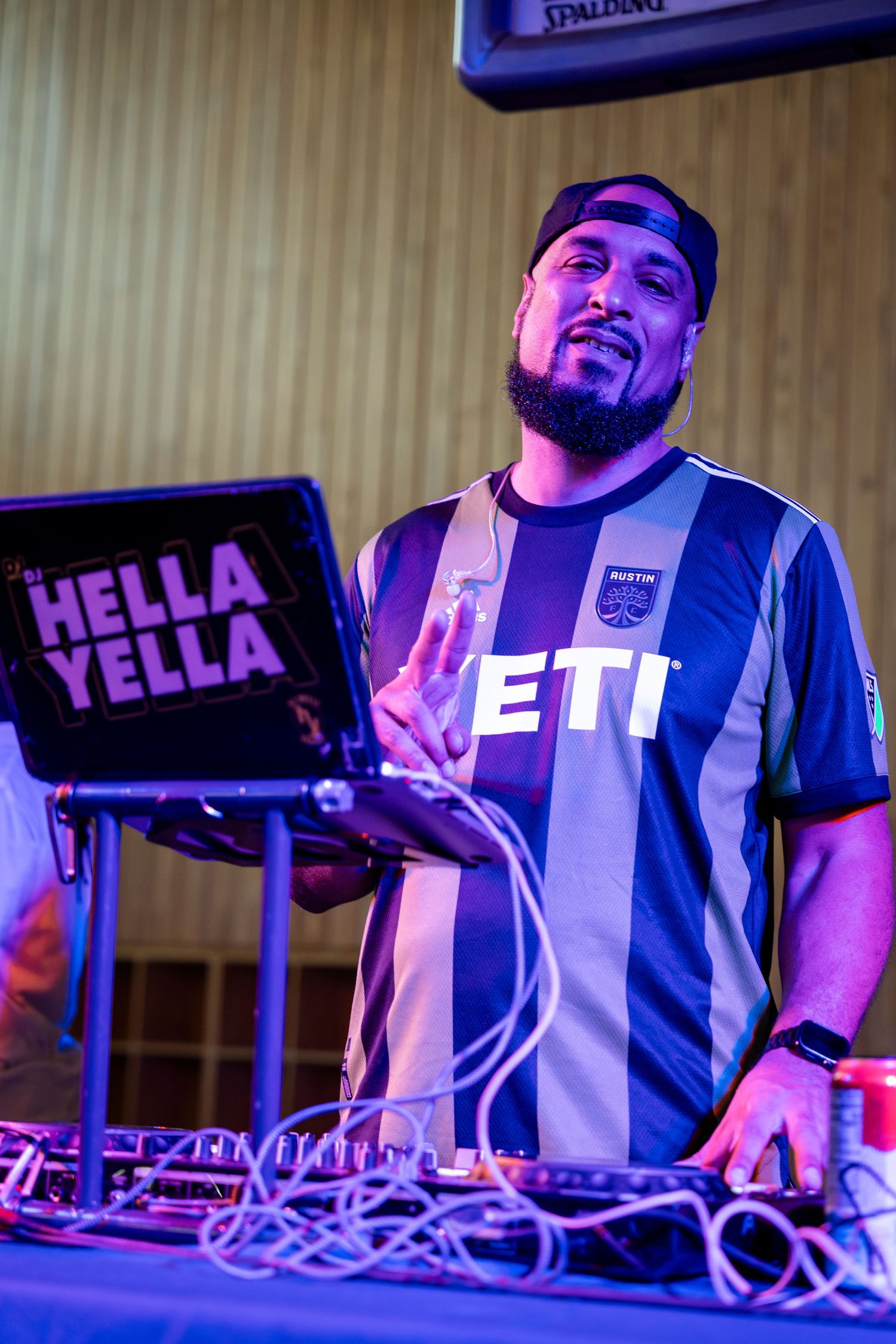 DJ performing at an event with purple lighting, wearing a striped jersey and standing behind equipment with a Hella Yella sign.