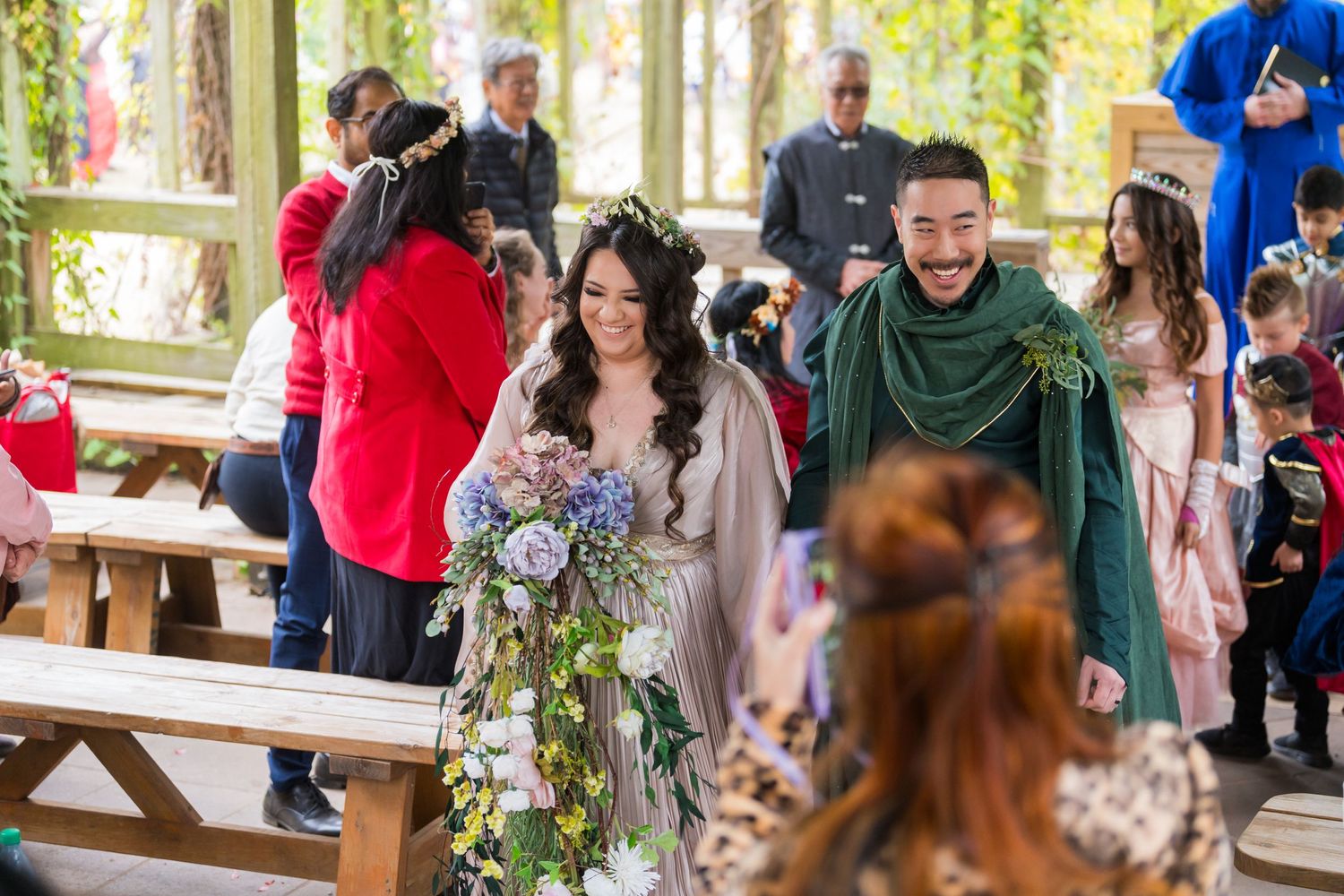 Wedding celebration with guests gathered around picnic tables under trees decorated with purple flowers.