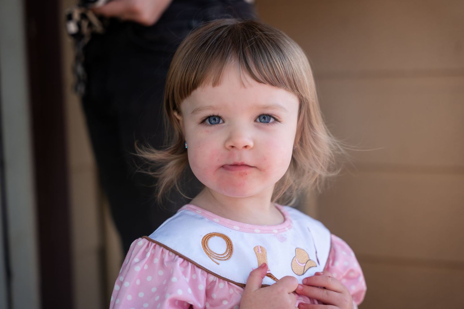 A young child wearing a pink and white dress looks at the camera with soft blonde hair and rosy cheeks.