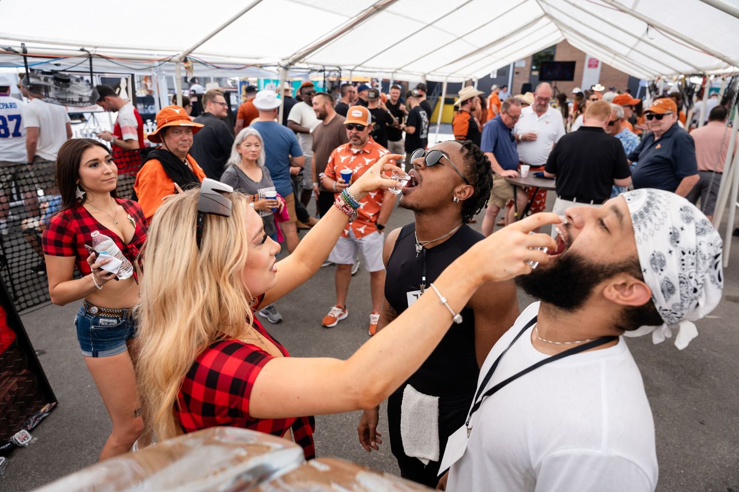 Partygoers taking shots at crowded outdoor event under white tent.