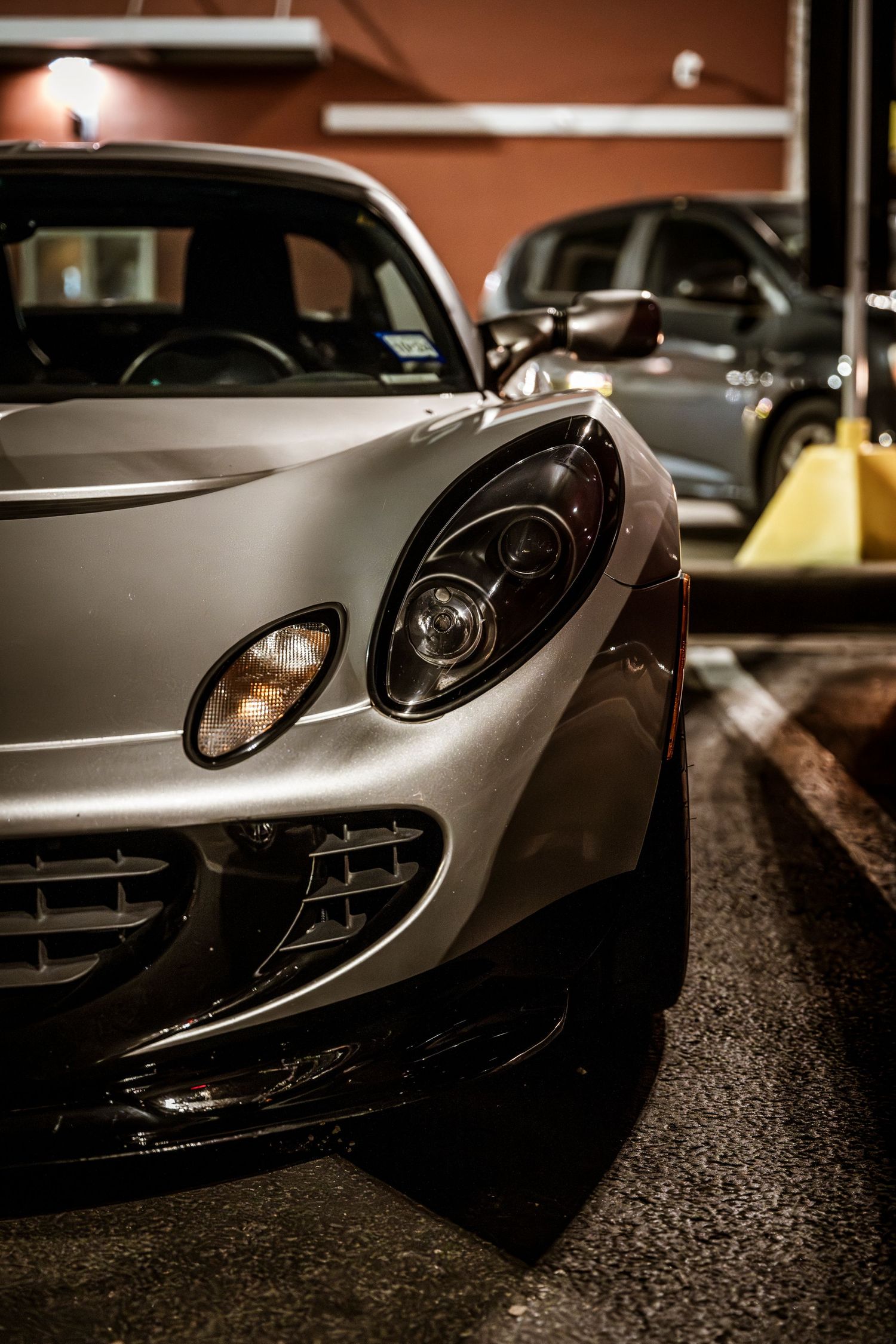 Close-up view of a silver Lotus sports car headlight and front quarter panel in dim lighting.