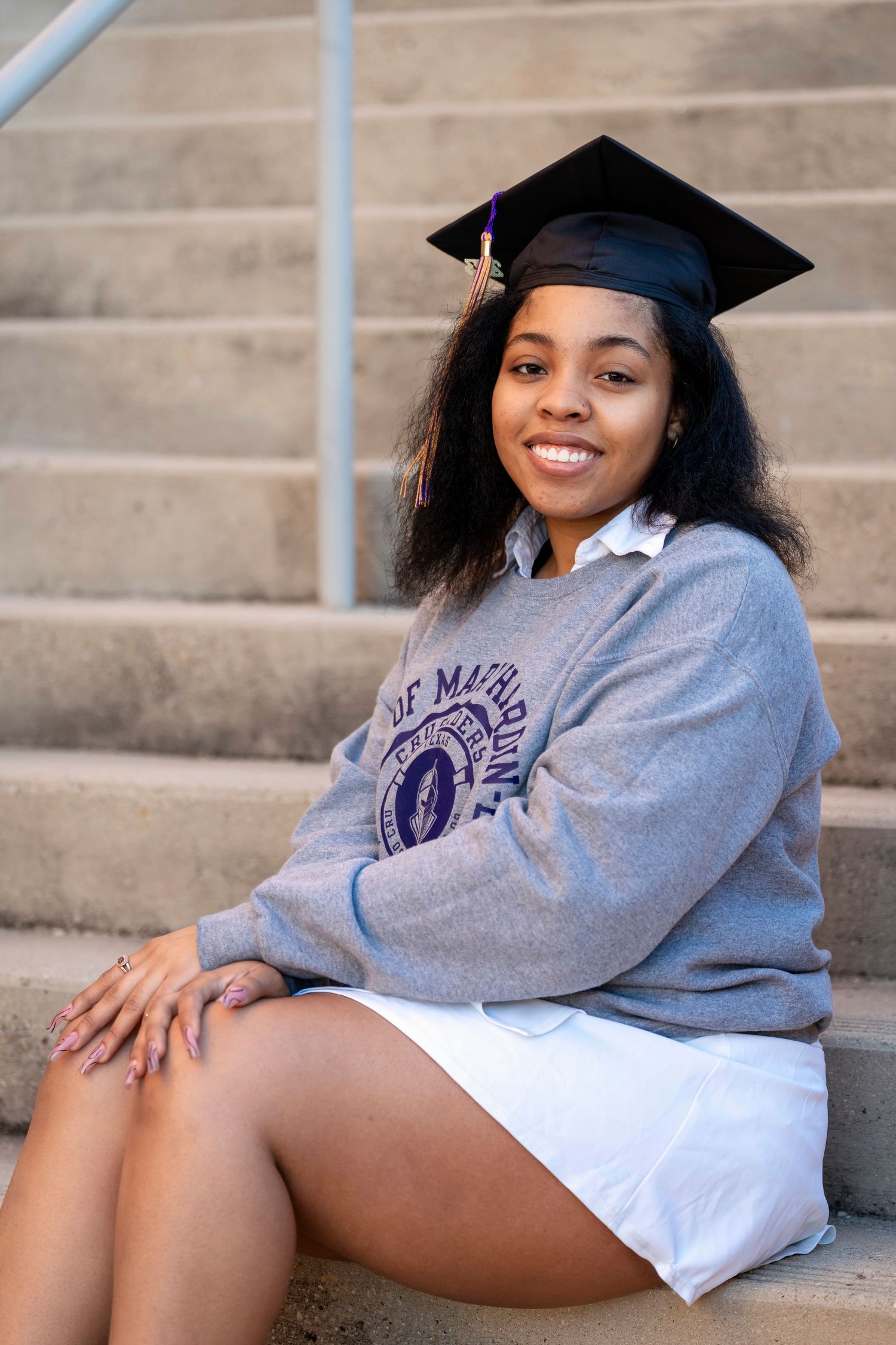 A graduate in academic regalia sits on concrete steps wearing a University of Mary Hardin Baylor sweatshirt.