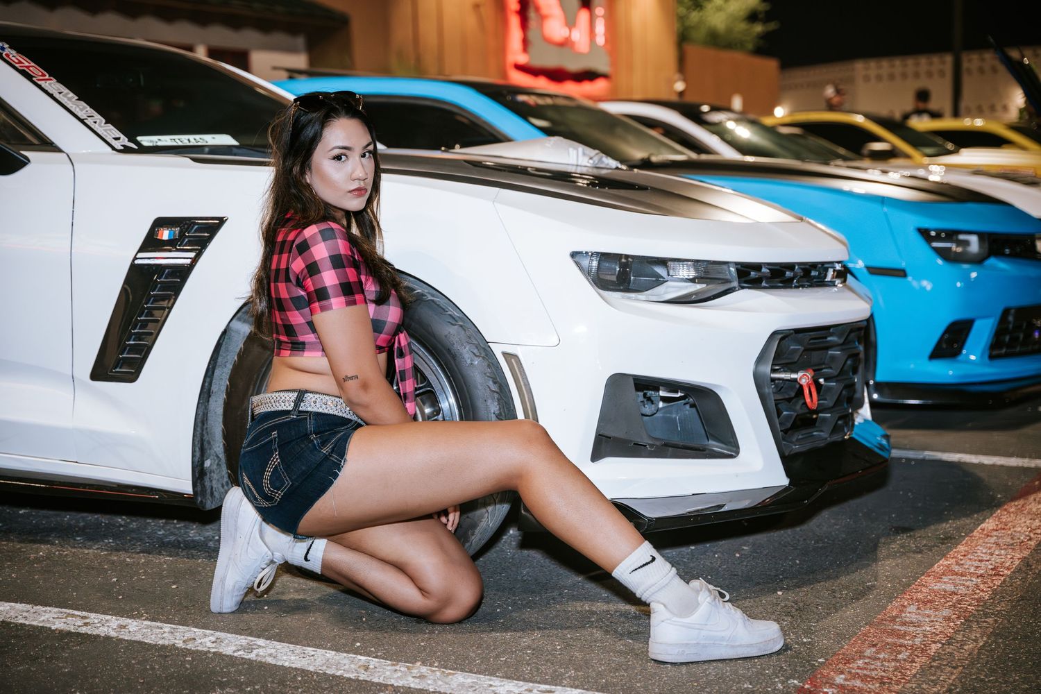 Model in plaid crop top crouches beside white sports car in parking lot.