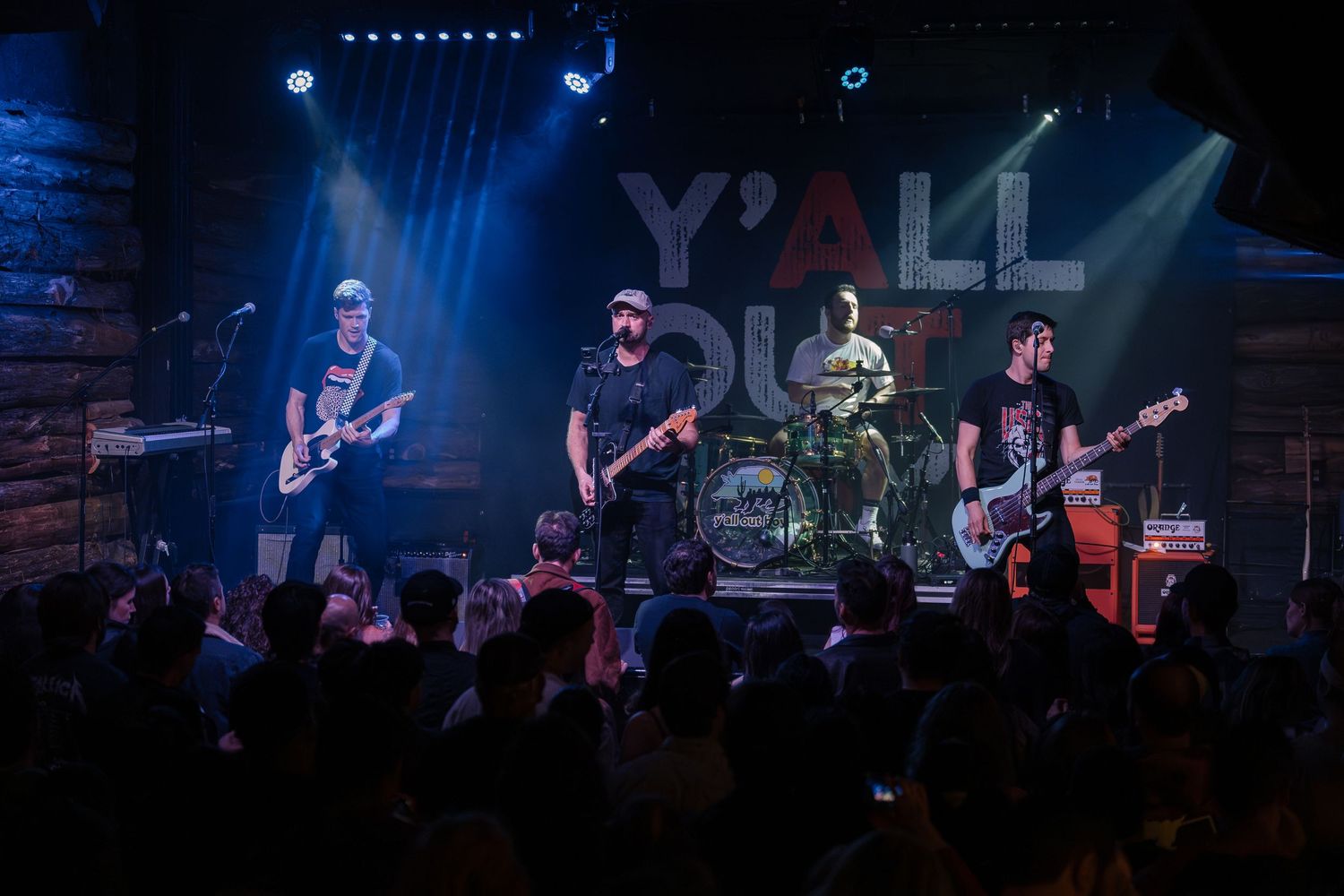 A full band performs on stage at a packed concert venue with professional blue lighting.