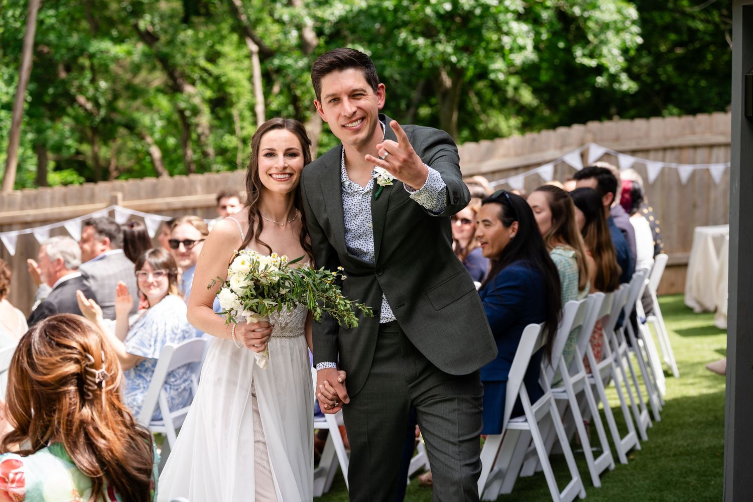 Joyful newlyweds exit their outdoor wedding ceremony while guests celebrate with white folding chairs visible.