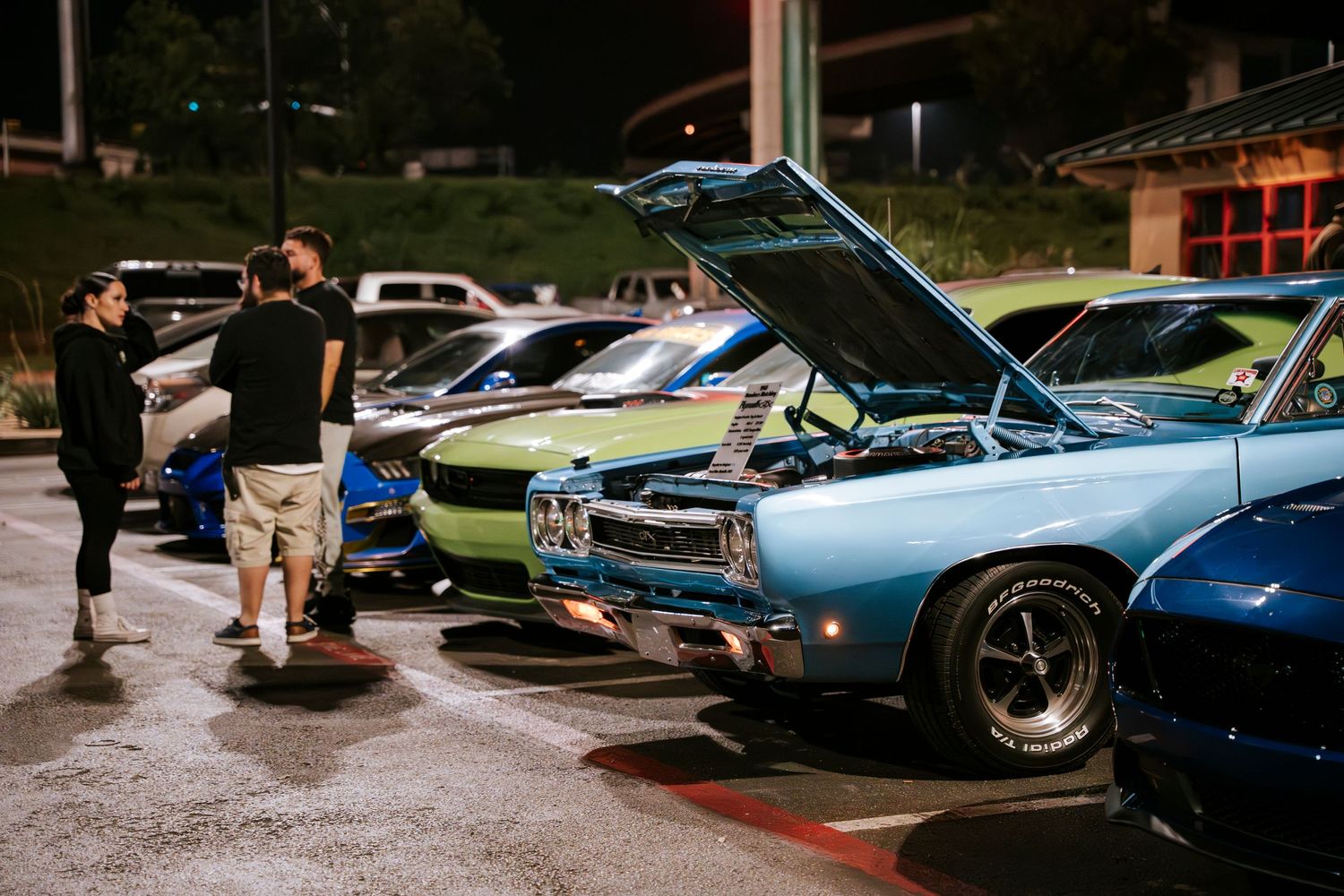 People examine classic muscle cars with open hoods at nighttime car meet.
