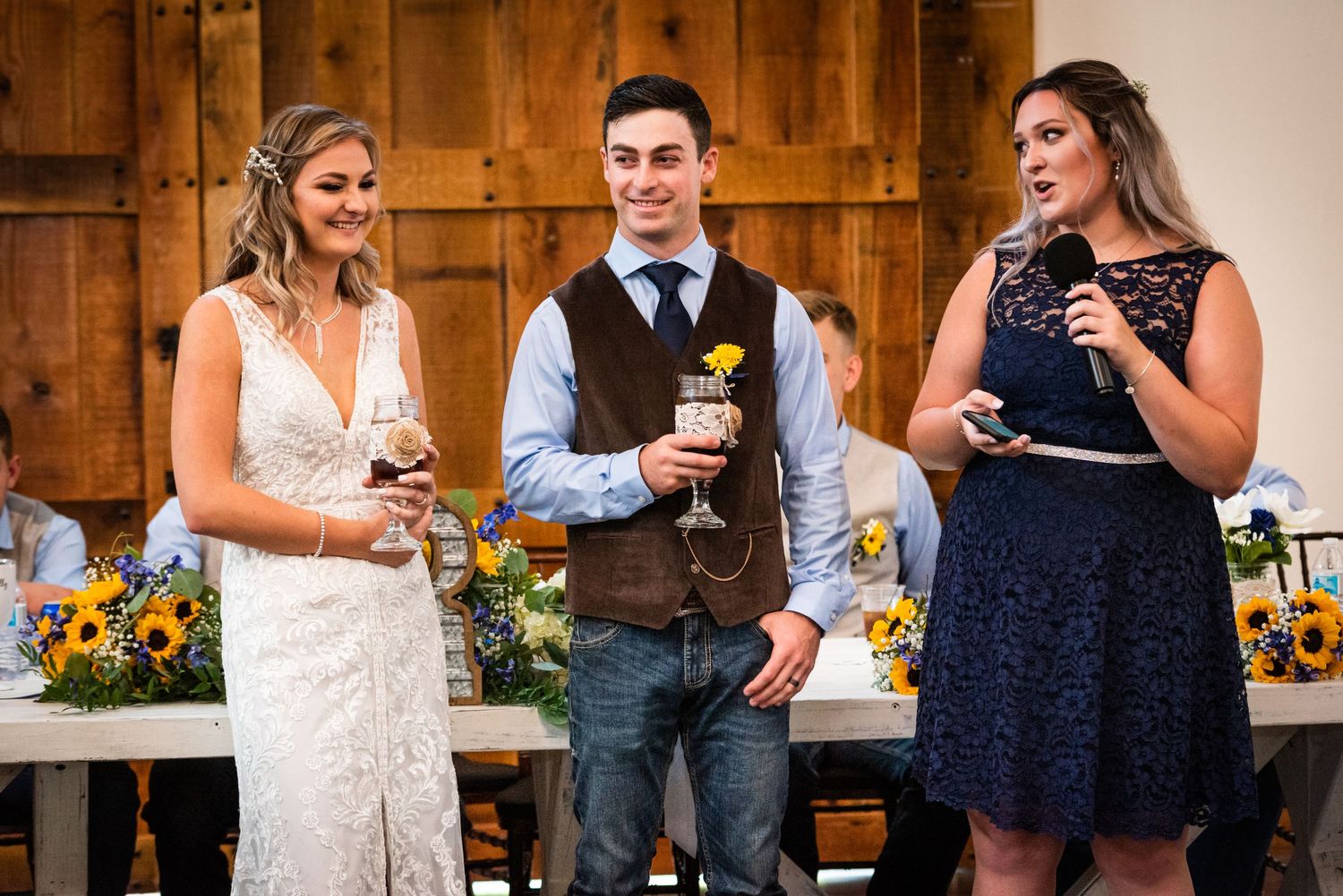 Wedding party members share toasts and laughs during reception speeches at a rustic venue.