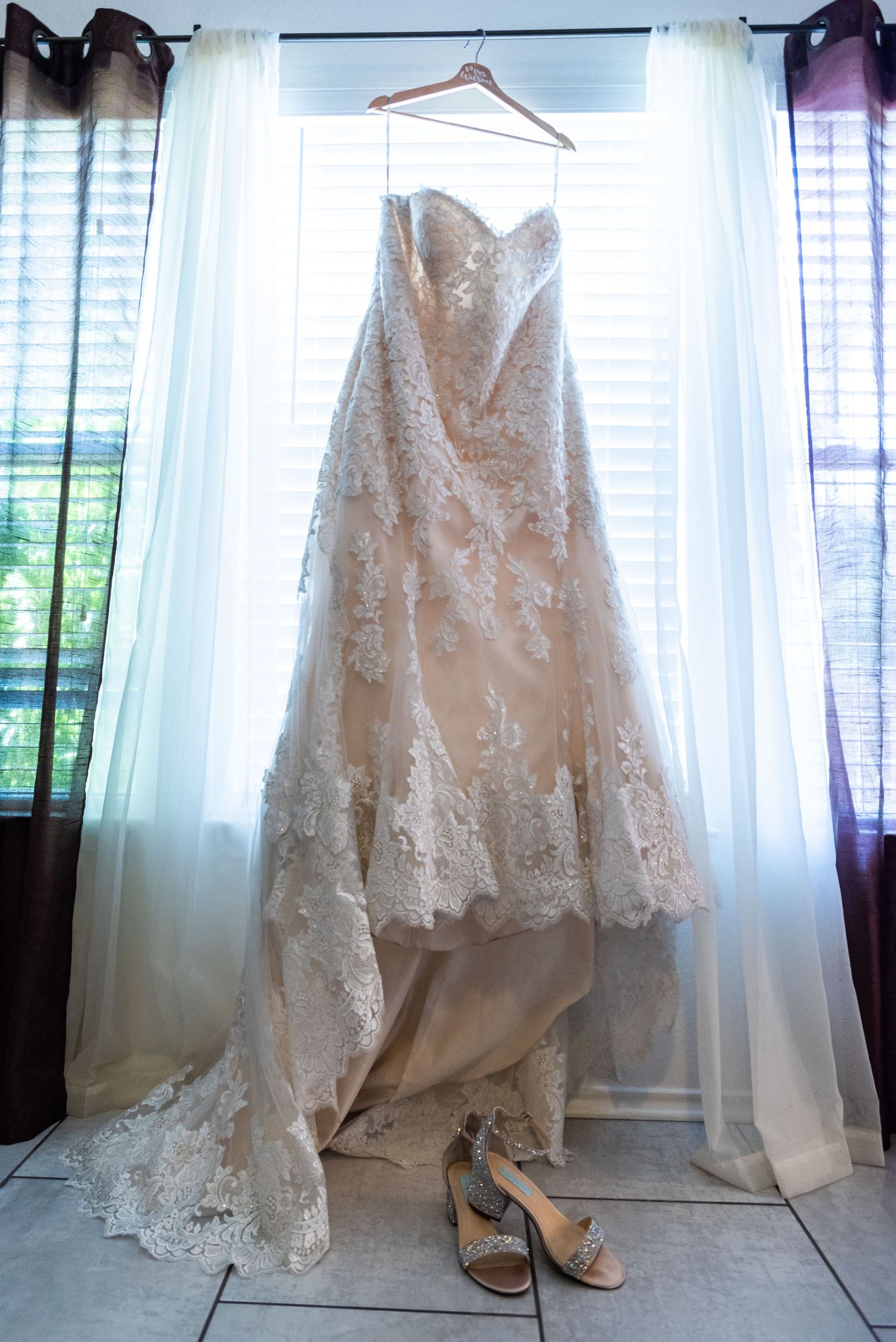 Elegant white lace wedding gown displayed with bridal shoes against window light.