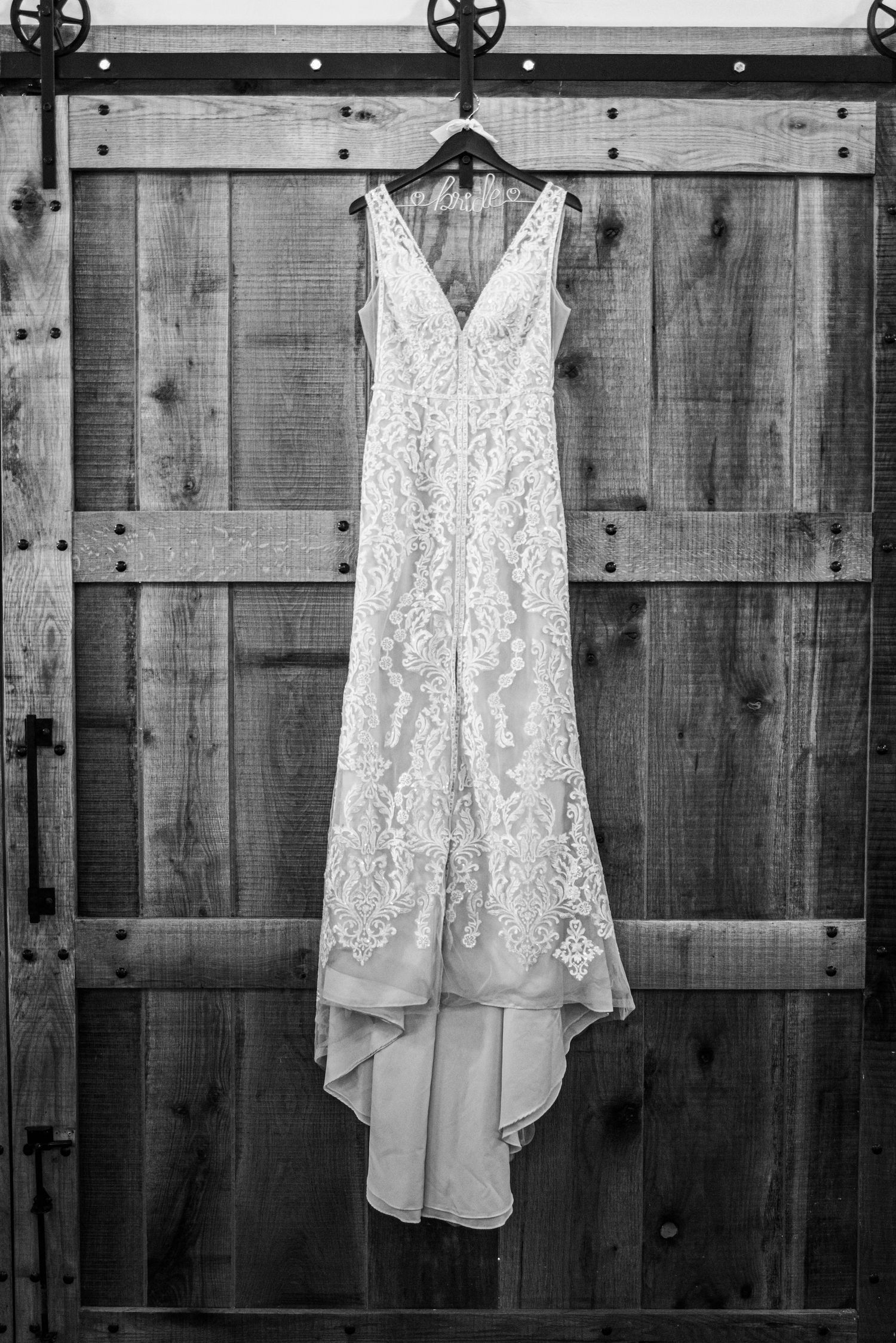A white lace wedding dress hangs on a rustic wooden door in black and white.