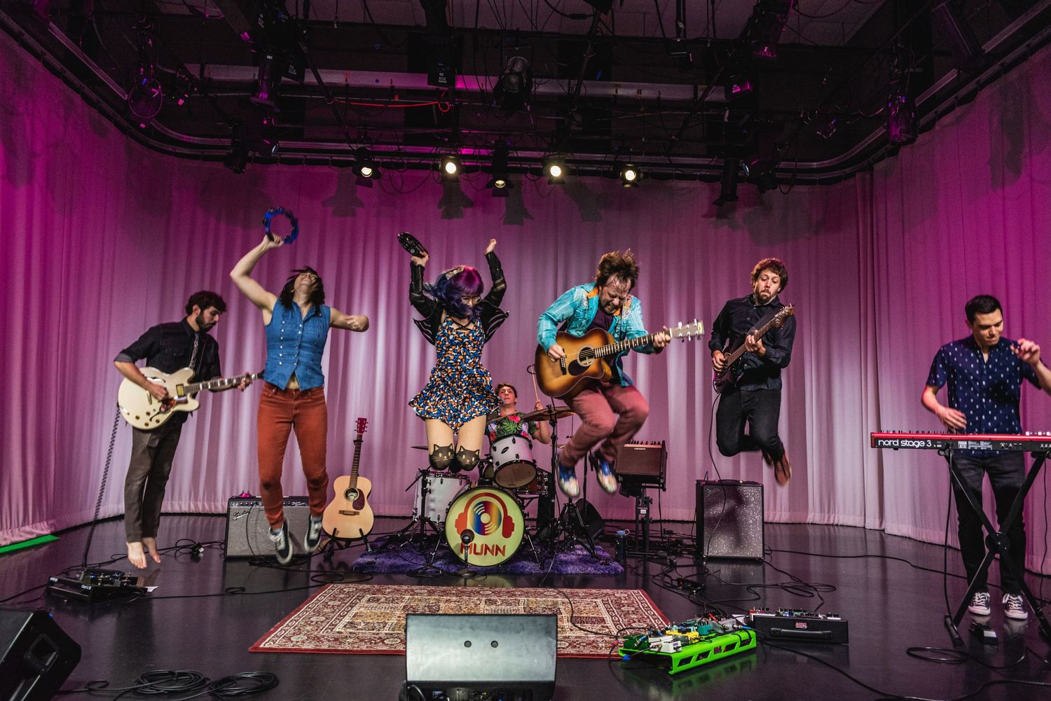 Musicians perform on stage with instruments and drum kit against magenta curtain backdrop.