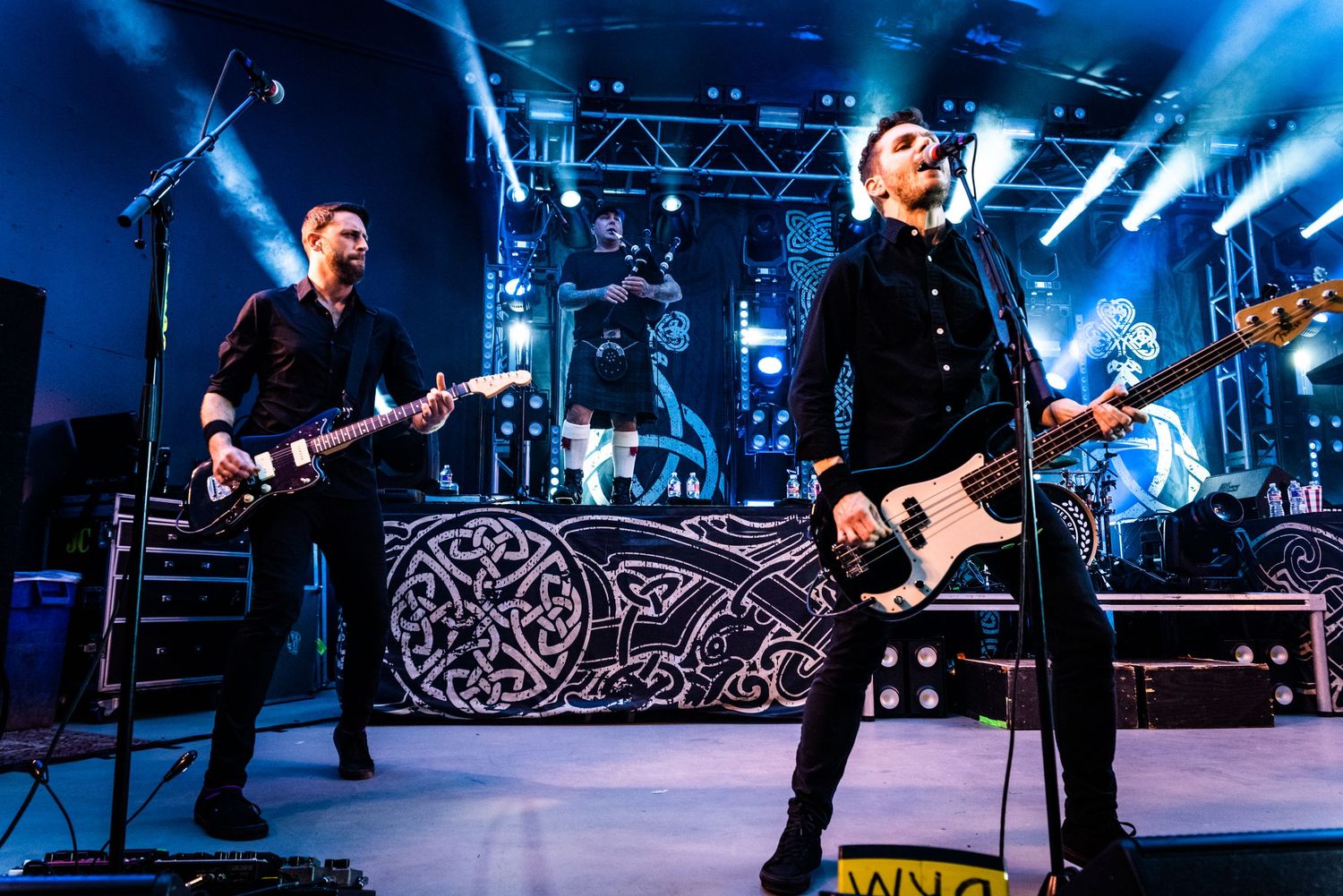Musicians perform with electric guitars on a concert stage with dramatic blue lighting and stage equipment.