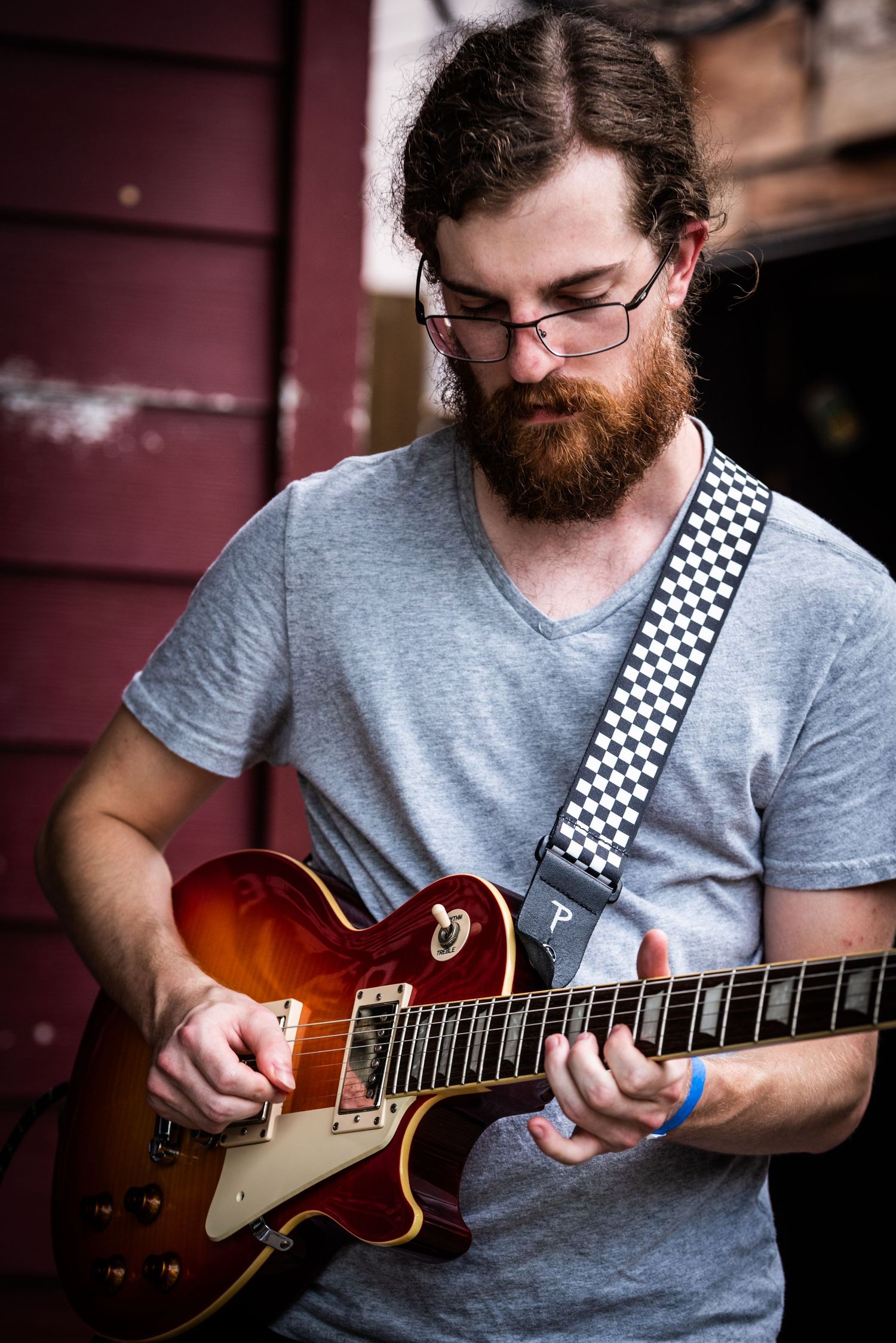 Guitarist in grey shirt with checkerboard strap focuses intently on playing Les Paul style guitar.